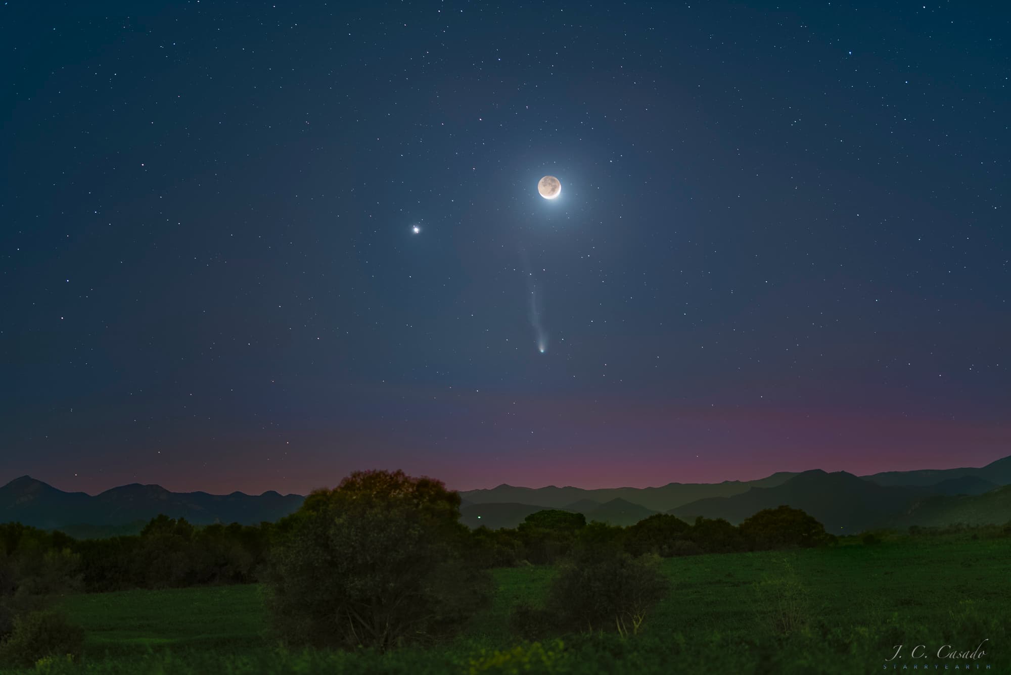 سماءٌ مُرصّعة بالنجوم فوق مشهدٍ أرضيٍّ داكنٍ معشوشب، تظهر فيها ثلاثة أجرام ساطعة، هي المُشتري في أعلى اليسار وقمرٌ هلال في أعلى اليمين والمُذنّب پونز-بروكس تحتهما حيث يصنعون مُثلّثاً. يُرى ذيلان يمتدّان من المُذنّب إلى الأعلى تقريباً.