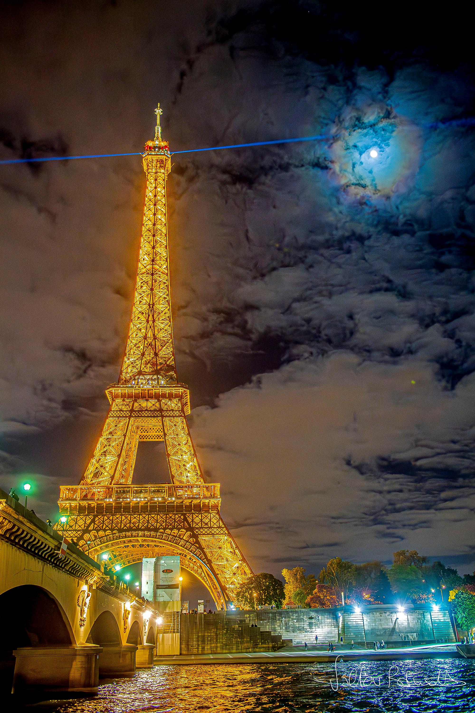 يظهر برج إيفِل الشهير في پاريس، فرنسا، يساراً مُناراً بالذهبيّ ليلاً. يُشعُّ ضوء ليزر أزرق للخارج من قمّته. تُرقِّط السُّحُب سماء الخلفيّة، ويكون القمر مرئيّاً أيضاً عبرها، لكنَّه مُحاطٌ بحلقاتٍ زاهية بالألوان: إكليلٌ قمريّ.