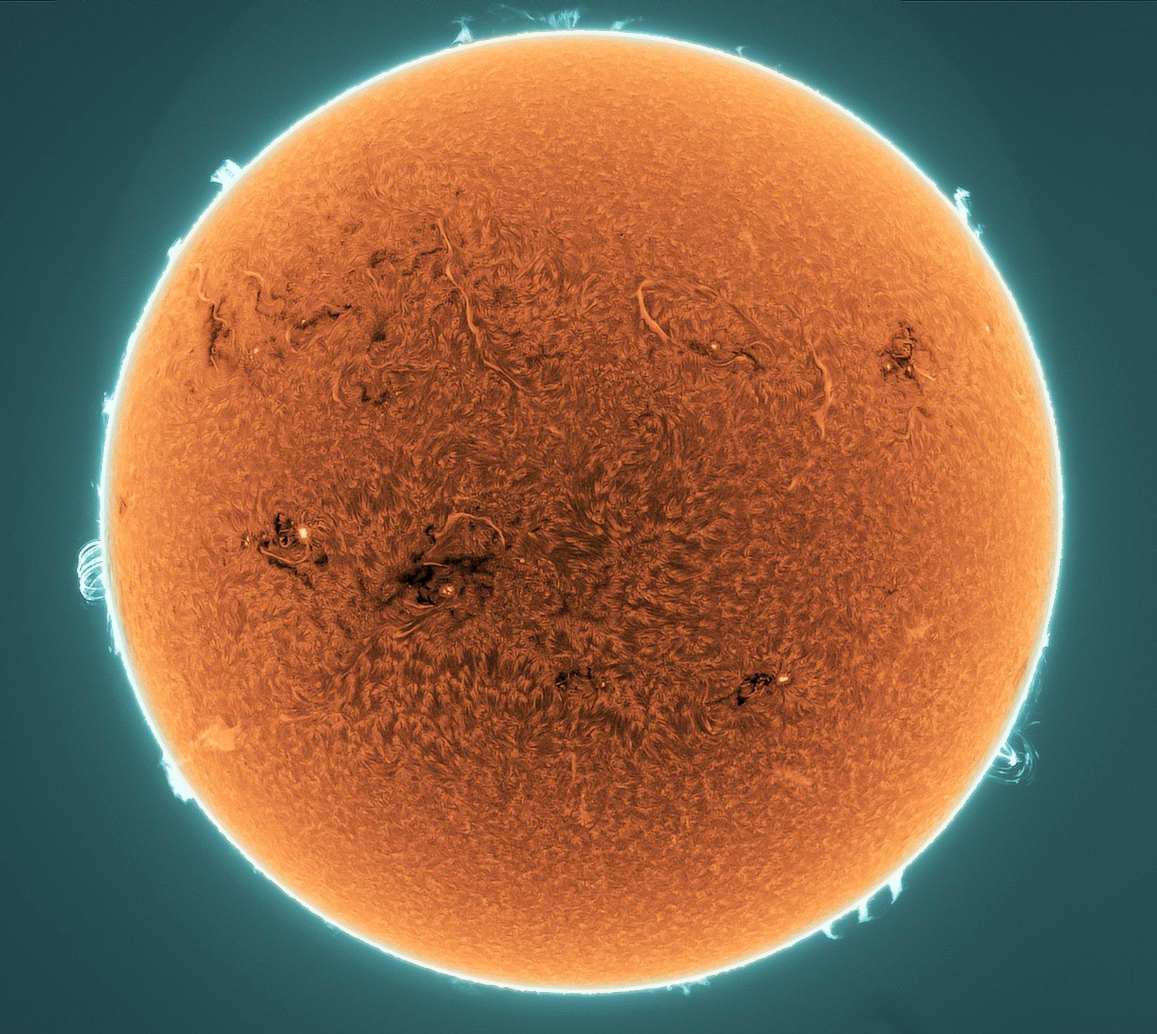 قرص الشمس بلون برتقالي يبدو وكأنّه مكسوٌّ بخيوط بعضها كبير في حين تظهر آثار النشاط الشمسي عند الحافّة على خلفيّةٍ مخضرّة