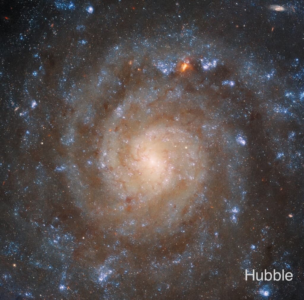 صورة هَبل تظهر المجرّة بالضوء المرئي بنواة مصفرة وأذرع ملتفة وممرات غبار معتمة بينها مع تناثر مناطق النجوم الزرقاء