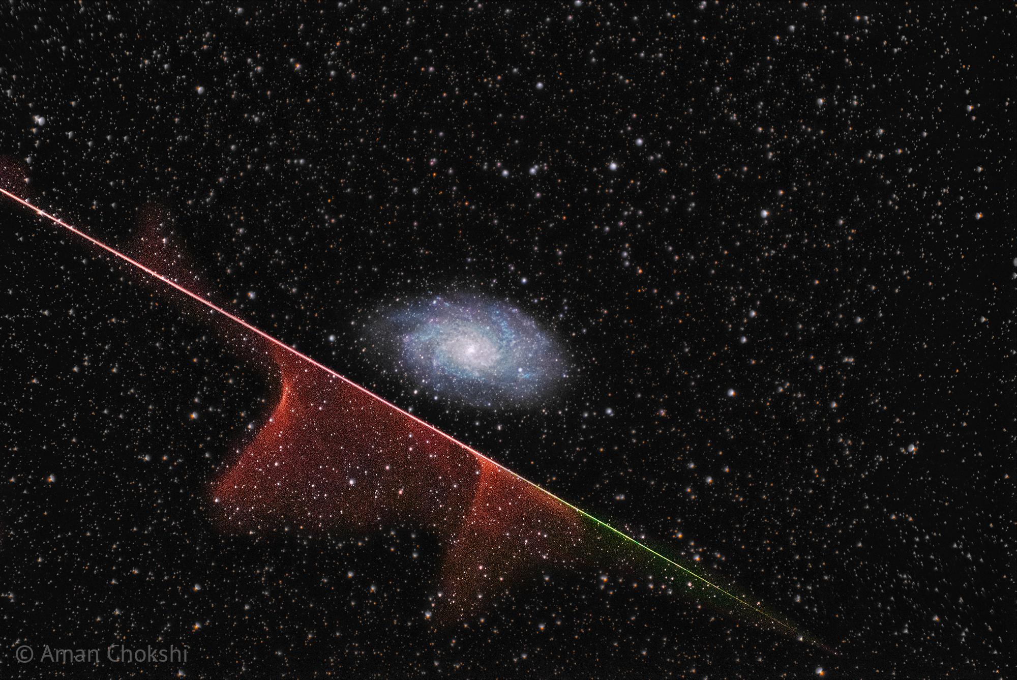 مجرّة حلزونيّة بعيدة في مركز الإطار يعبر تحتها من أعلى اليسار إلى أسفل اليمين خطٌّ مُتعدّد الألوان تظهر أغبرة مُتلاشية قرب بعض أجزائه. تتناثر النجوم عبر الصورة.