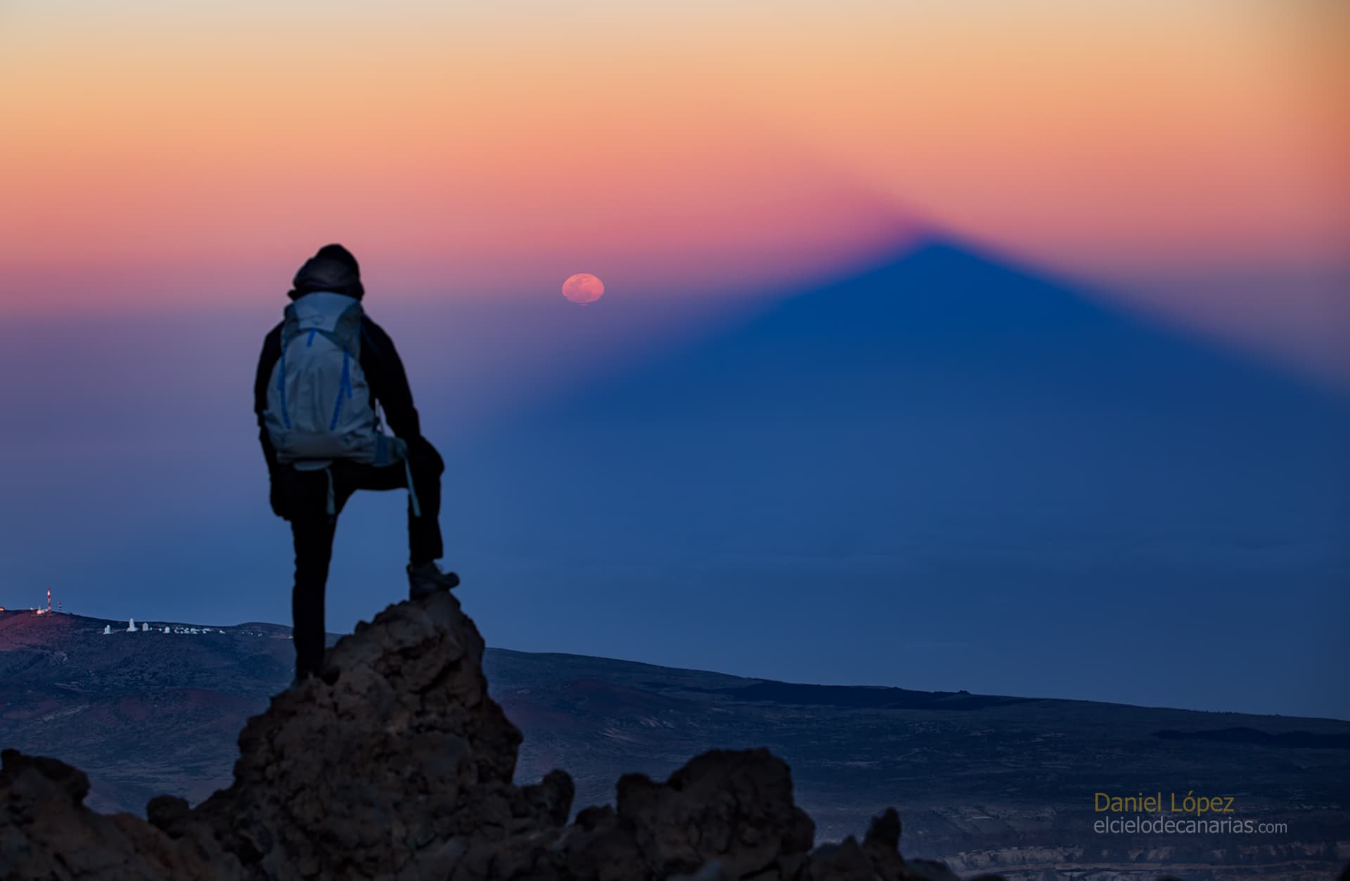 شخصٌ على صخور ينظر أماماً باتّجاه أُفقٍ يظهر فيه مُثلّثّ ظلٍّ داكن وعلى ميسرته بدرٌ ساطعٌ قرب الأُفق