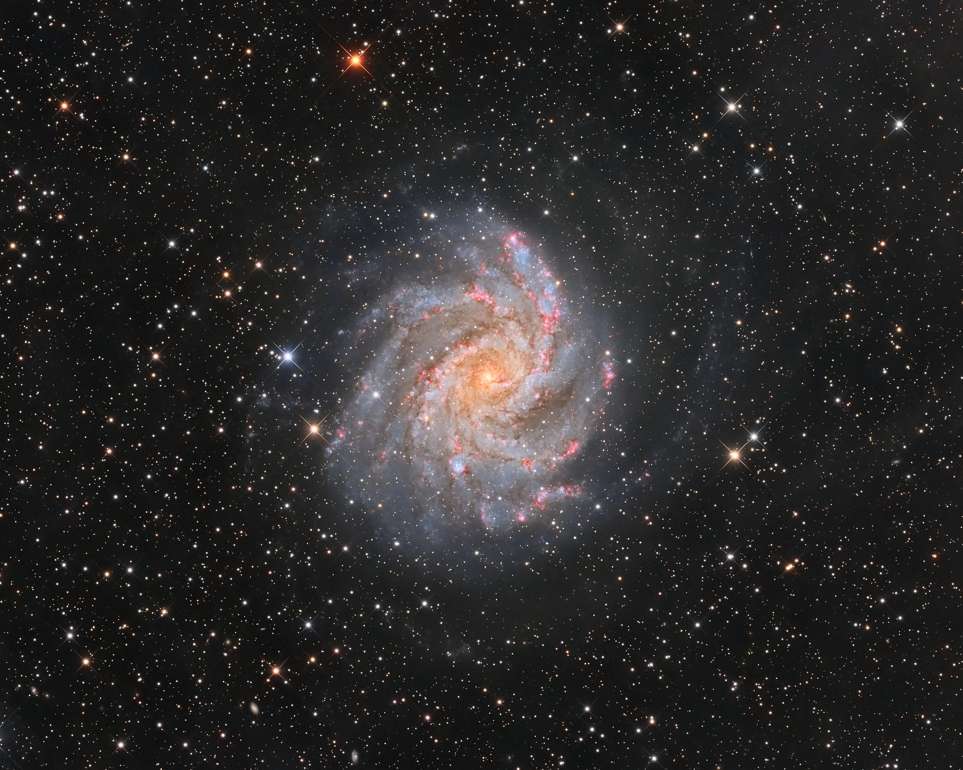 مجرّة حلزونيّة تُرى من وجهها بنواتها المصفرّة التي تمتدّ منها أذرع مزرقّة تشوبها مناطق محمرّة في حين تتناثر نجوم المقدّمة عبر الصورة