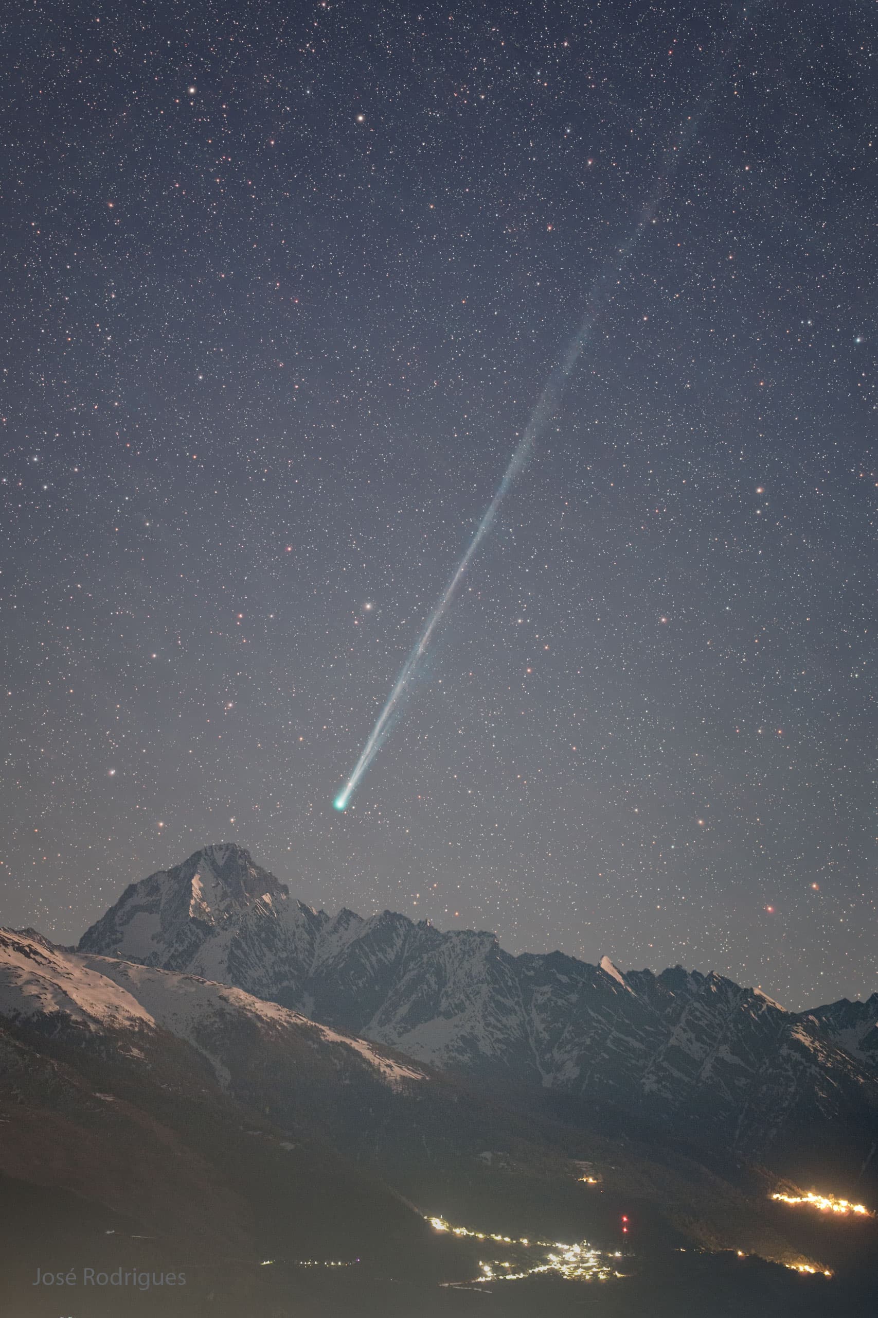 ليلة مرصّعة بالنجوم فوق جبال في المقدّمة. باتّجاه اليمين يوجد مذنّب رأسه بالقرب من أسفل المركز، مع ذيل طويل يمتدّ باتّجاه أعلى اليمين.