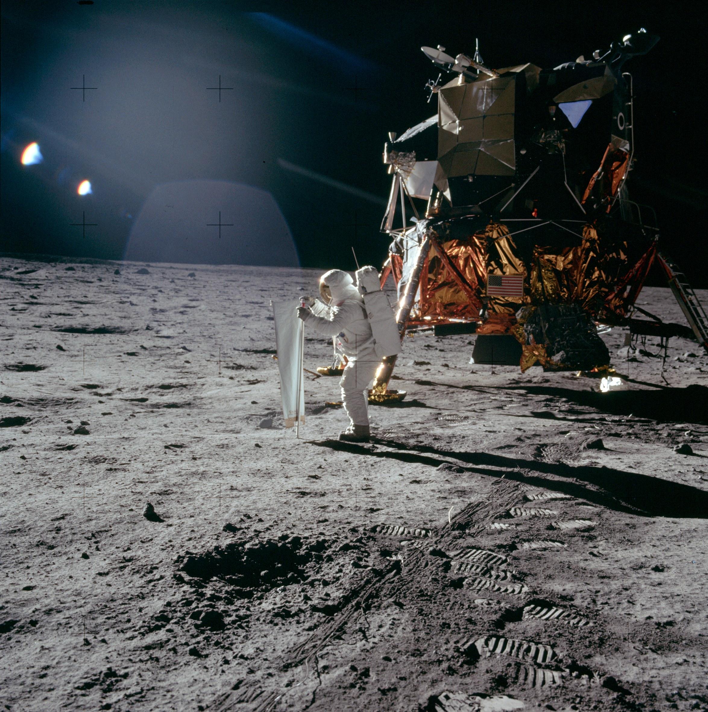 رائد فضاء ينشر طبقة بينما تلمع أشعّة الشمس. تظهر خلفه وحدة قمريّة في حين تظهر أثار دعسات الأحذية القمريّة تحته.