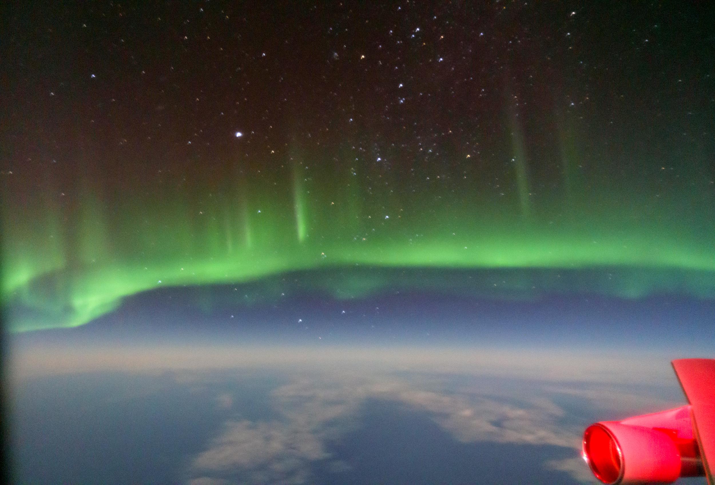 شفق قطبي أخضر وفوقه السماء بتجومها وتحته جزء منها أيضاً والأرض، ويظهر طرف طائرة على مايبدو بلون أحمر أسفل اليمين