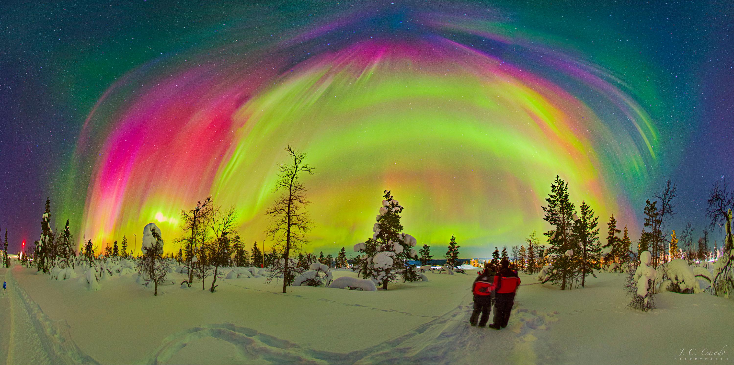 يقف شخصان يرتديان معاطف حمراء على مشهدٍ طبيعي ثلجي به أشجار عارية. في الأعلى، تظهر العديد من الأشفاق القطبيّة بألوان مختلفة ، مع بعض النجوم المرئيّة في الخلفية.