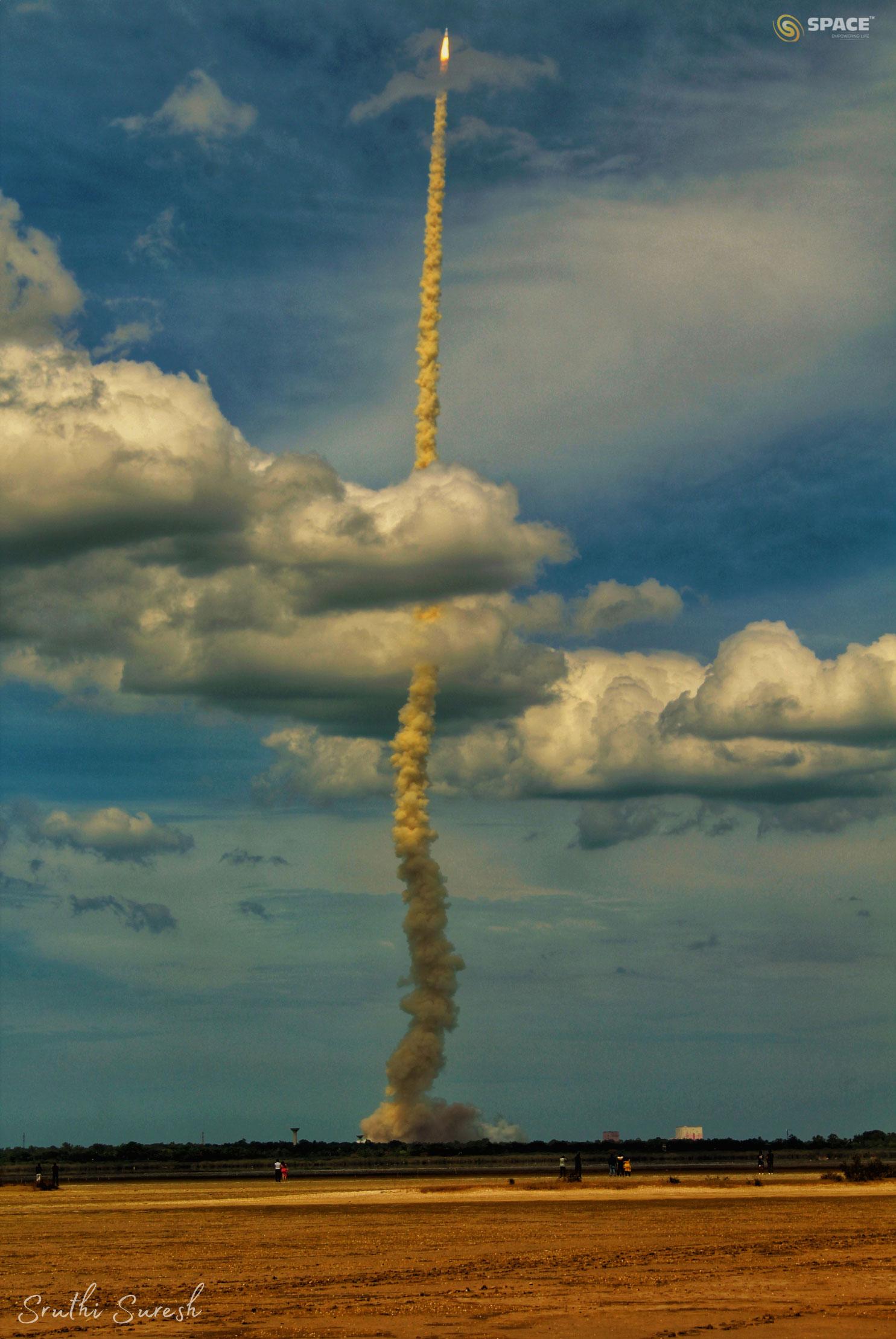 يُرى صاروخٌ بعد الإقلاع بعامود دخّانٍ طويل. التُقِطَ الصاروخ قبالة سماءٍ زرقاء وقد عبر من خلال مجموعة سُحُب. في المُقدِّمة يوجد حقلٌ فارغٌ داكن اللون.
