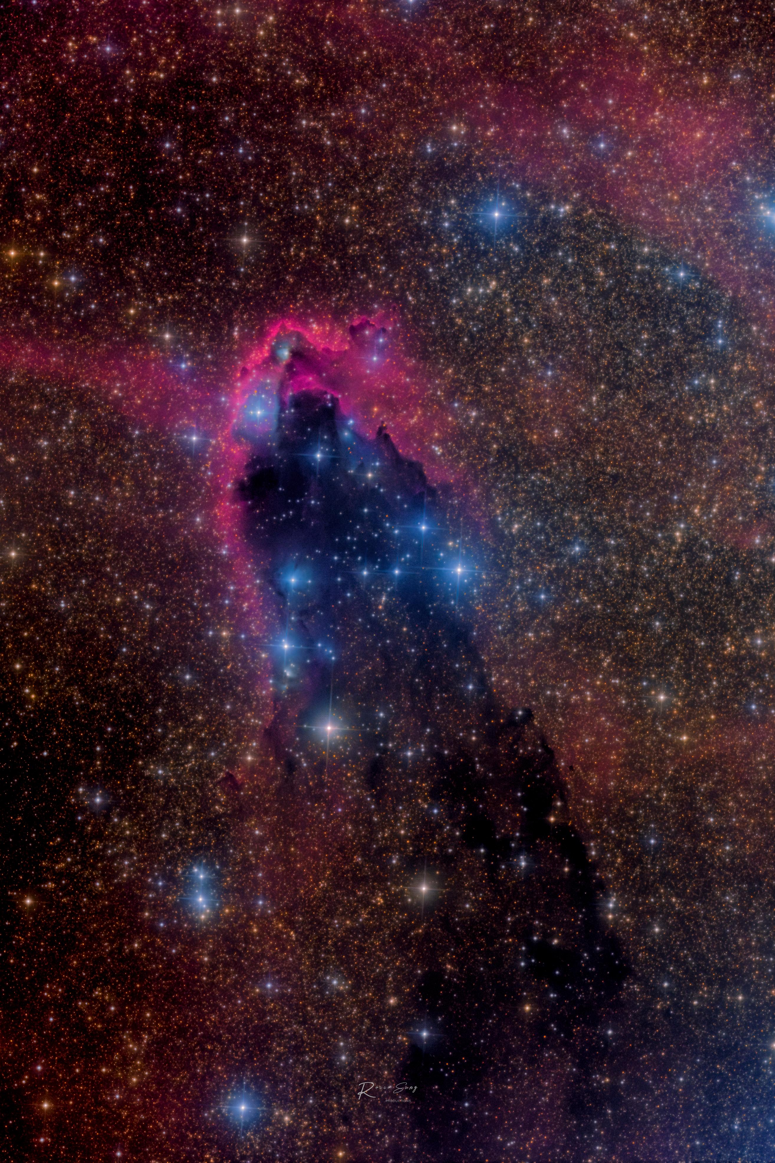 صورة مزدانة بالنجوم للفضاء العميق يظهر في وسطها امتداد داكن فيه نجوم زرقاء يحاذيها من الأعلى غازات ذات وهج محمرّ.