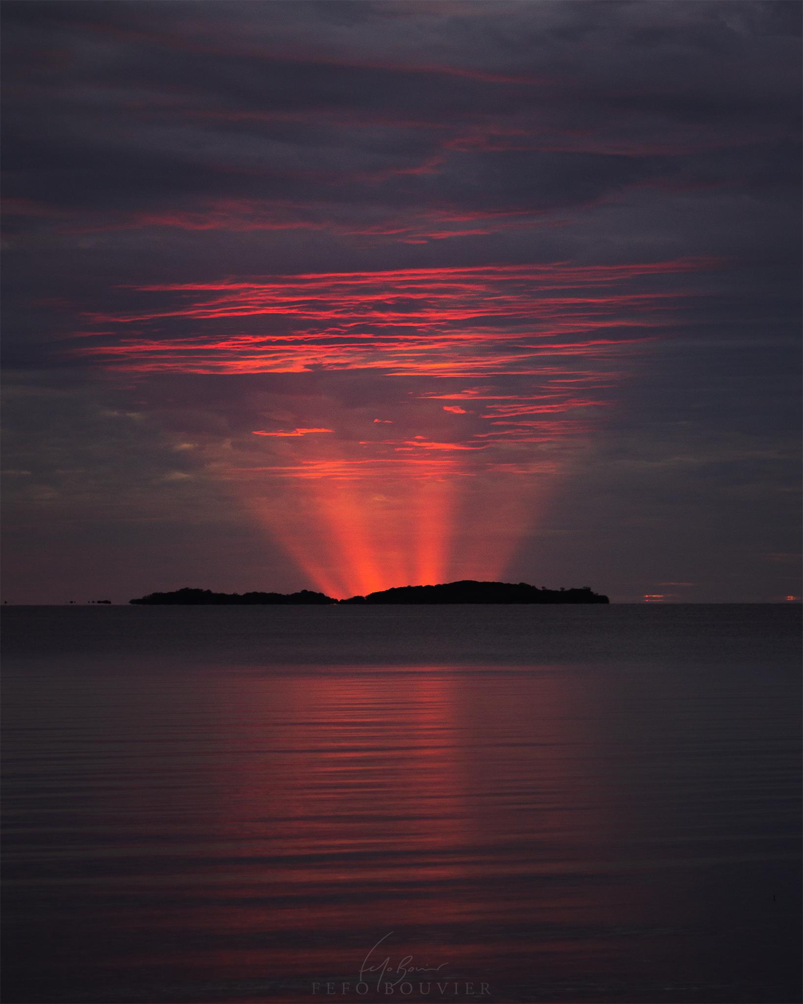 جزيرة بعيدة ينبثق ورائها من نقطة تحت الأفق أشعّة حمراء تنعكس على الغيوم في أعلى السماء ومنها على سطح الماء الذي أمامها في المقدّمة