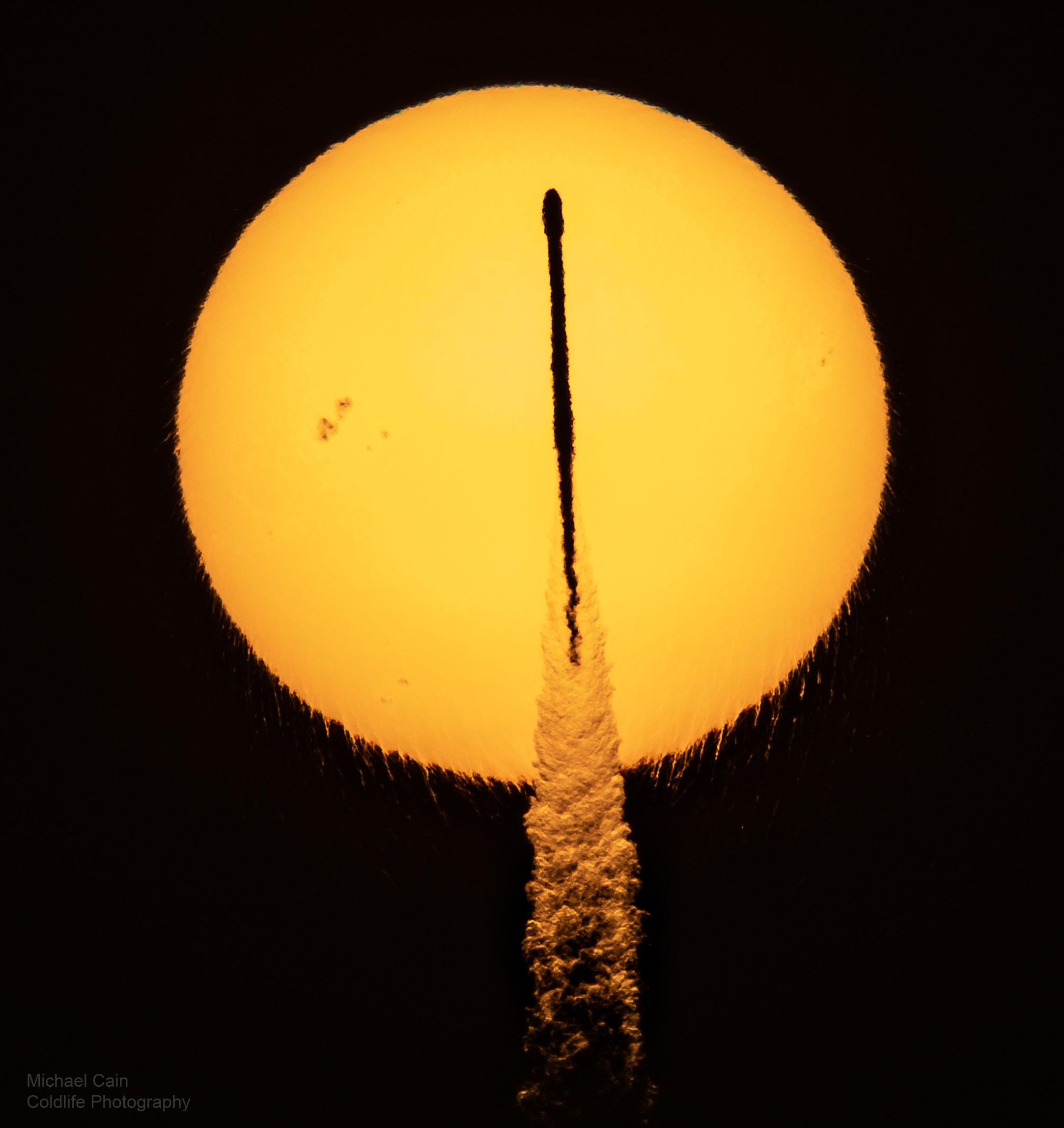 قرص الشمس وأمامه ظَليل صاروخ يظهر كعود ثقاب متعرج قليلاً وتحته عمود دخانه. تظهر الشمس متموجة في الأسفل كذلك مع عبور بقعة شمسية في يسارها