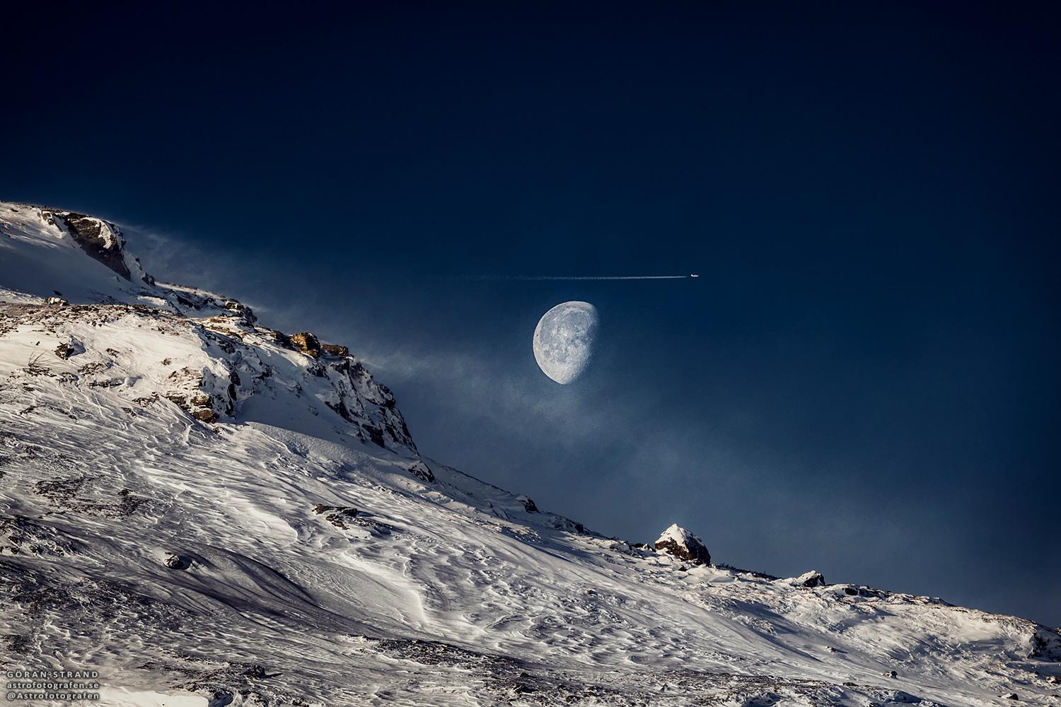 قمرٌ بدرٌ تقريباً يُرى فوق تلّةٍ مُنحدرة مكسوّة بالثلوج. تُرى طائرة ومسار تكاثف قُرب القمر مُباشرة.