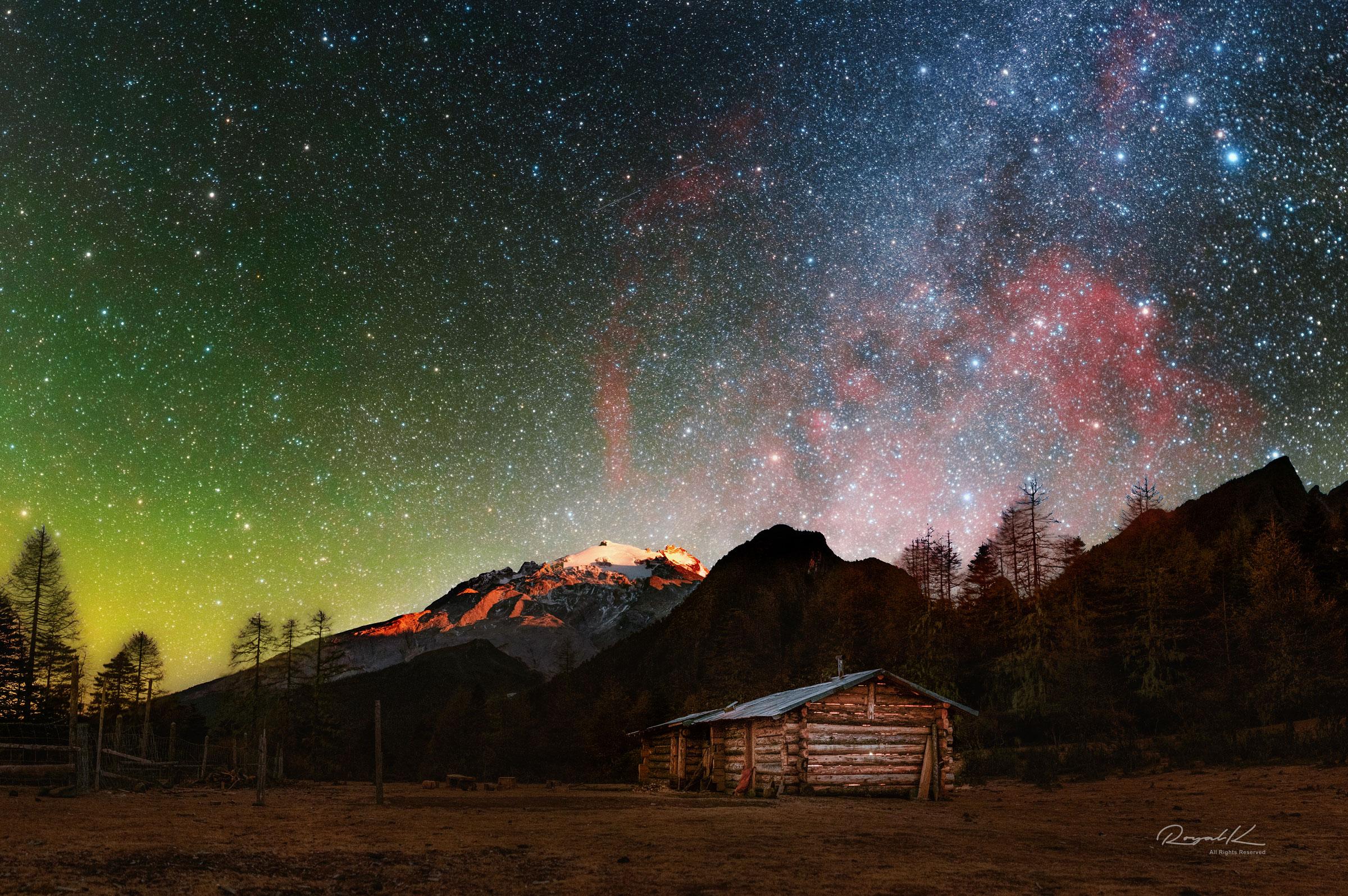 سماء ملوّنة يظهر فيها السديم المحمرّ الكبير وحوله النجوم وتحته جبل مكسو بالثلوج وفي المقدّمة أرض فيها كوخ خشبي وحوله أشجار