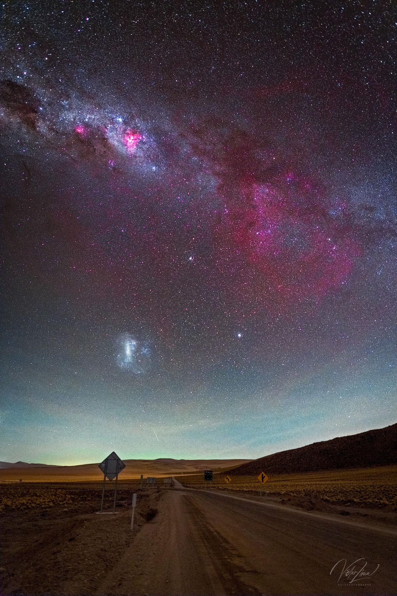 مشهدٌ سماويٌّ شاسع مع طريق صحراء بُنيّ في المُقدِّمة وسماءٍ تحتوي على نطاق درب التبّانة المجرّي بكامله مع وهجٍ أحمر كبير على اليمين وهو سديم "گام" الخافت, مجرّة (LMC) مرئيّةٌ أيضاً.