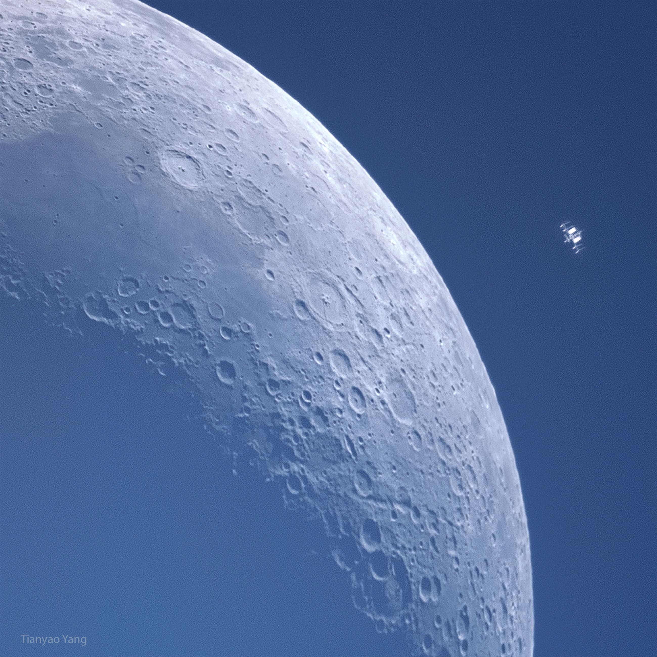 قمرٌ هلال على خلفيّةٍ زرقاء. تظهر الكثير من الفوّهات بتفاصيل عظيمة. يظهر إلى أعلى اليسار آلةٌ صغيرة من نوعٍ ما هي في الحقيقة محطّة الفضاء الدوليّة في مدارٍ حول الأرض أيضاً.