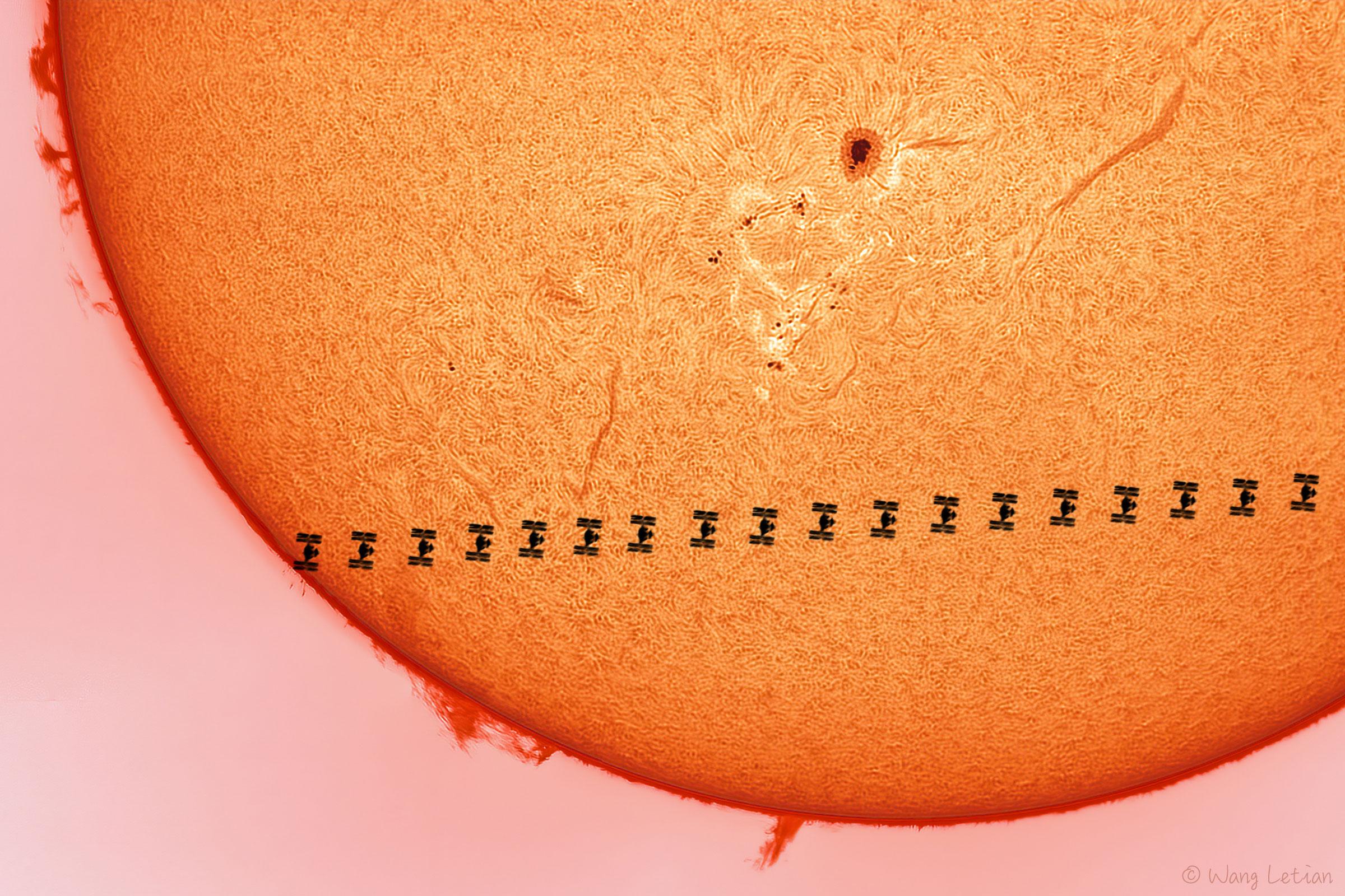 ثلث قرص الشمس تقريباً تعبر أمامه محطّة الفضاء الدولية بلقطات متقطّعة متتابعة بينما تظهر على سطحه الخيوط وبقعة شمسيّة داكنة وحوله الأشوظة