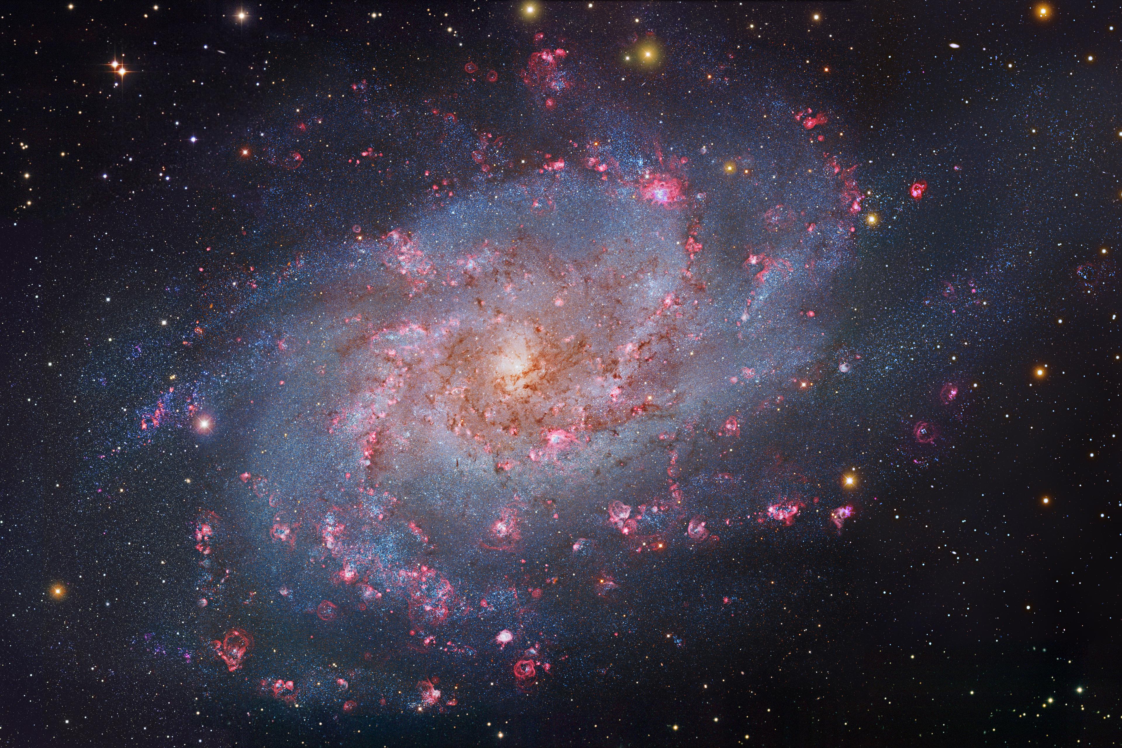 مجرّة تُرى من وجهها بأذرع حلزونيّة واسعة تحوي عناقيد نجميّة زرقاء ومناطق تشكّل نجميّ ورديّة
