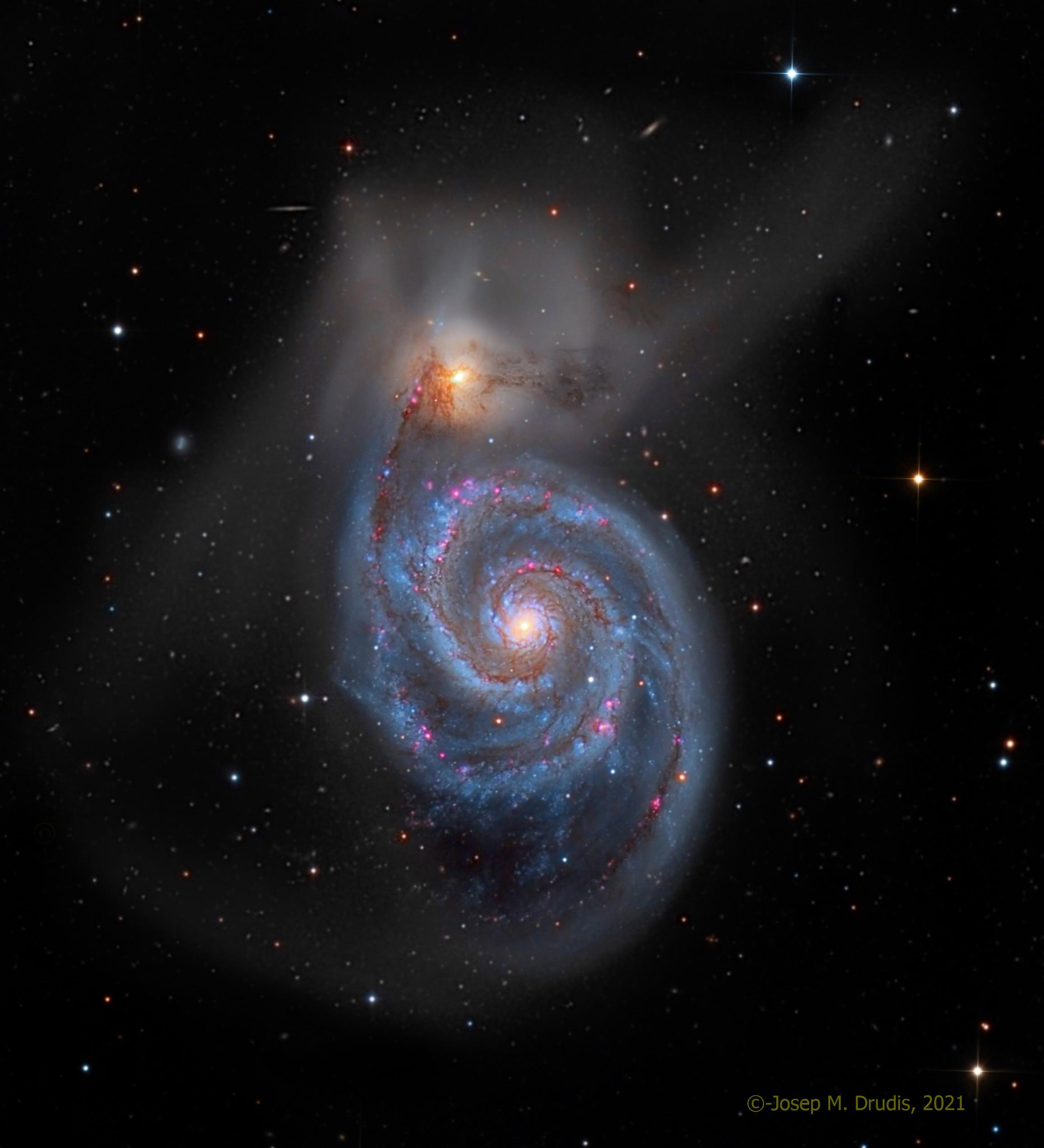 صورة للفضاء العميق تظهر مجرّة حلزونيّة وفي أعلاها مجرّة مصاحبة لها تبدو أصغر حجماً، بينما تتناثر أجرامٌ أخرى حولهما.