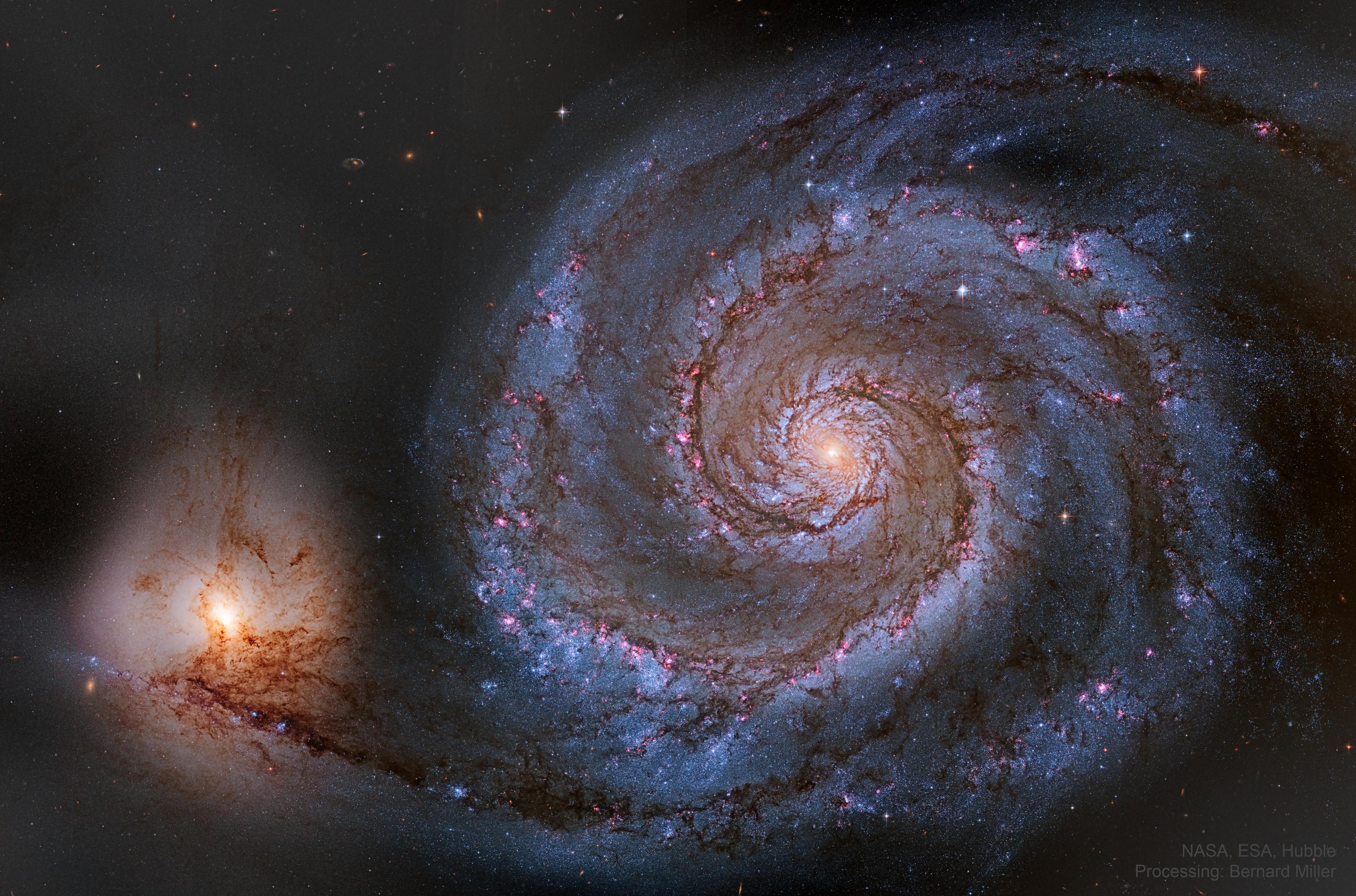 مجرّة حلزونيّة تُرى من وجهها بأذرعها التي تؤوي مناطق تشكل نجمي ورديّة ونواتها الصفراء الساطعة وعلى أسفل اليسار مجرّة أصغر متفاعلة ثقاليّاً يمتد إليها ذراع من المجرّة الكبيرة
