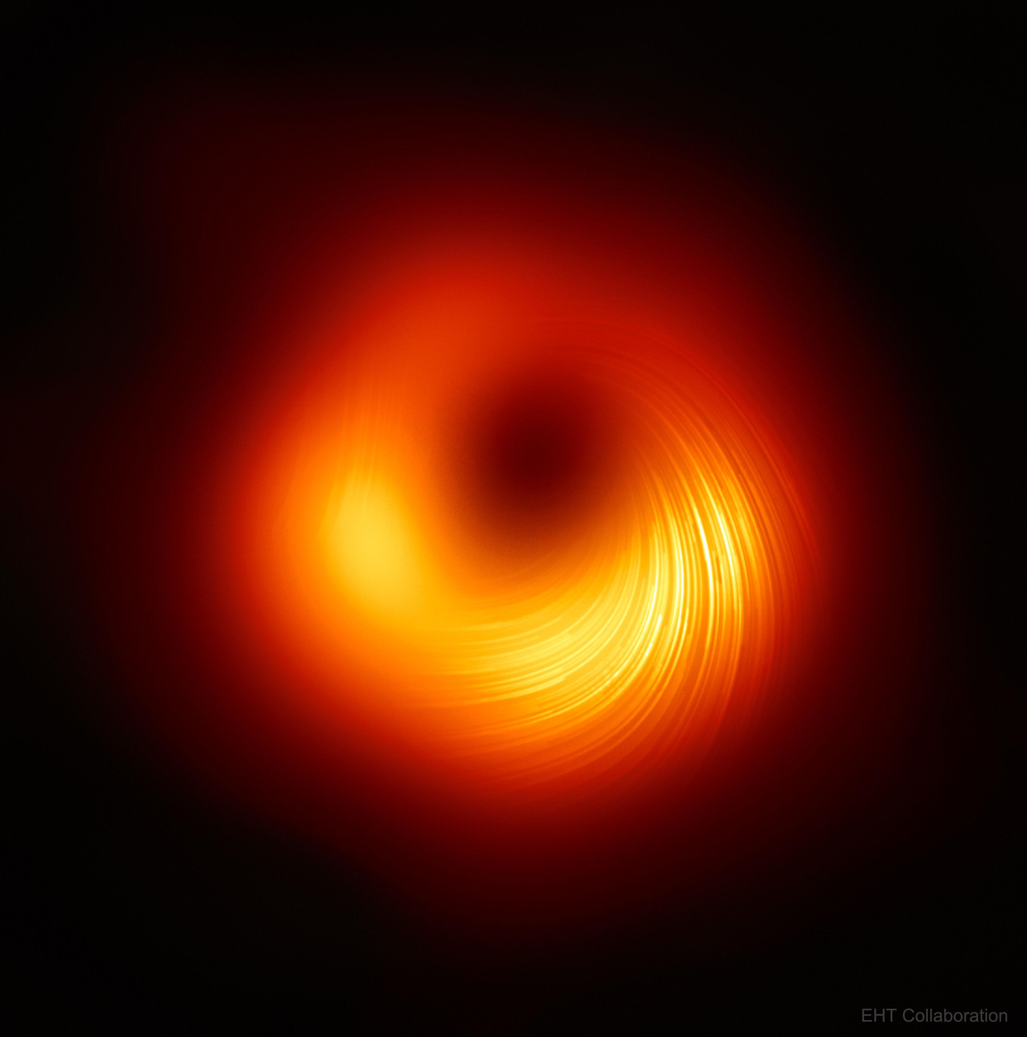 صورة للثقب الأسود في سماء سوداء، تظهر حلقة مضيئة محزّزة ذات ألوان متدرّجة بين الأصفر والأحمر تدور حول مركز مظلم.