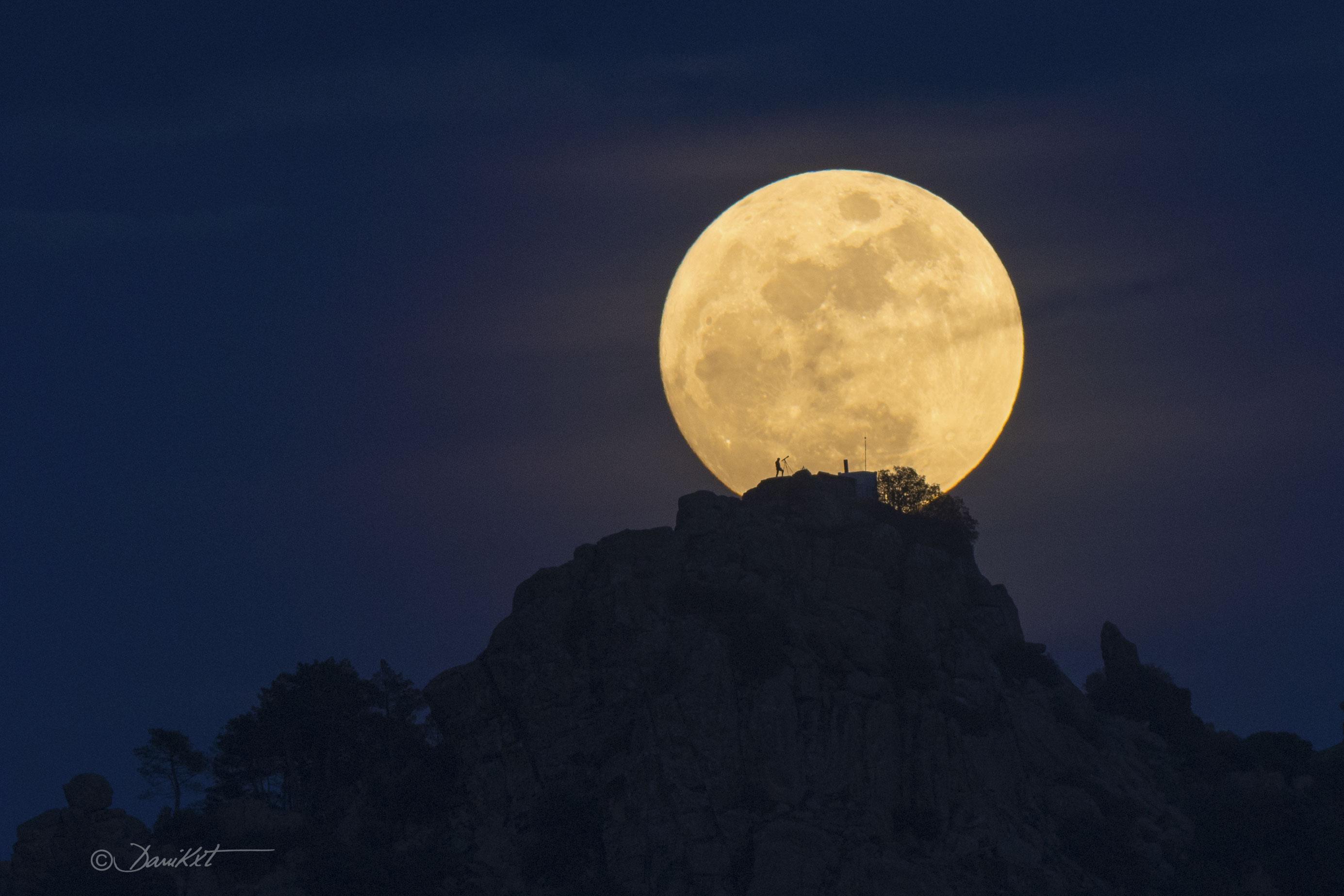 بدرٌ مُقرّب وأمامه مرتفع صخري عليه شخص مع تلسكوبه يظهر ك ظليل ومن خلفه القرص القمري الأصفر