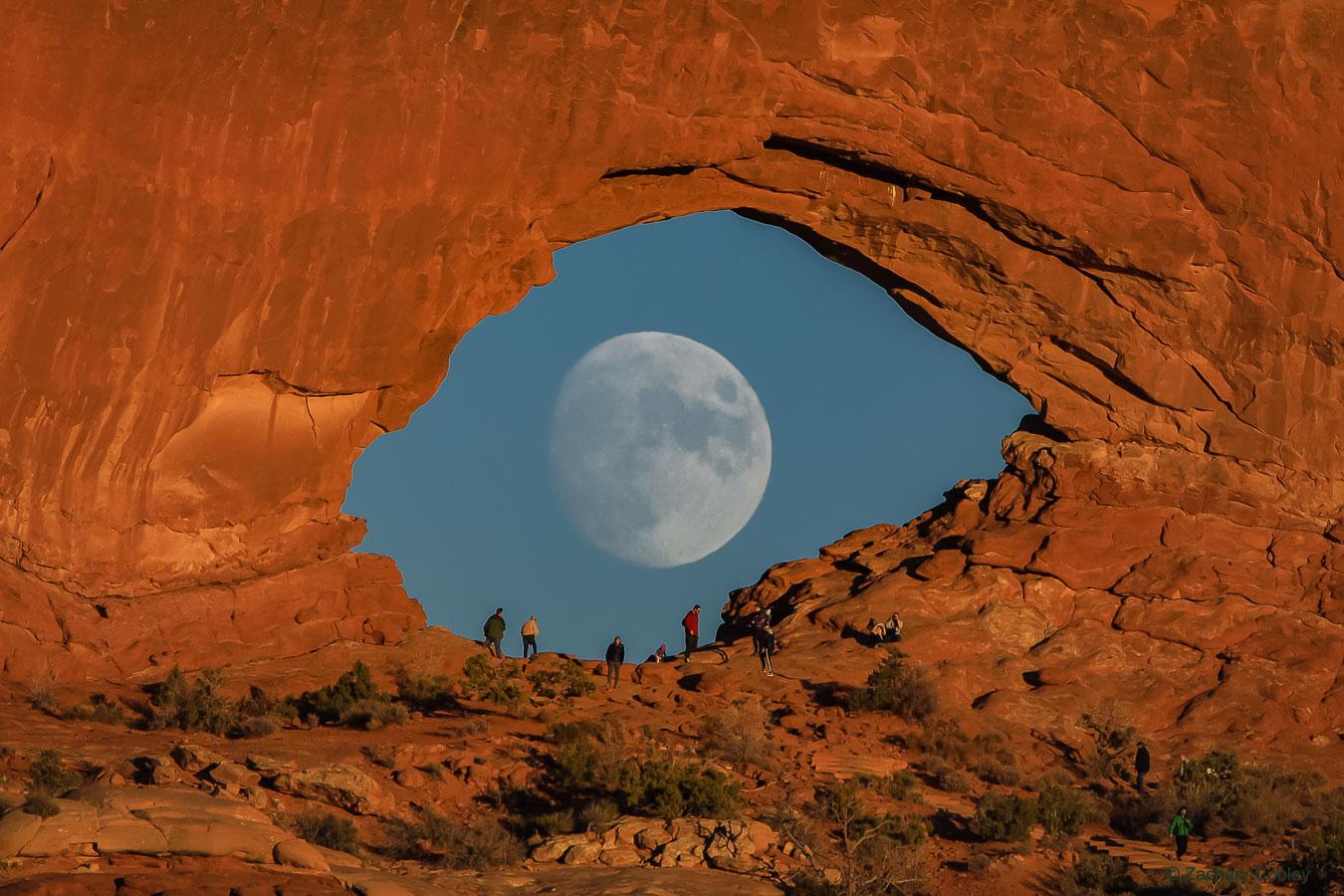 صورة للقمر عبر فتحة في حائط صخري (قوس الحجر الرملي) تبدو كأنها عين عملاقة تنظر بدورها إليك. هناك بعض الناس على الأرض تحت القوس