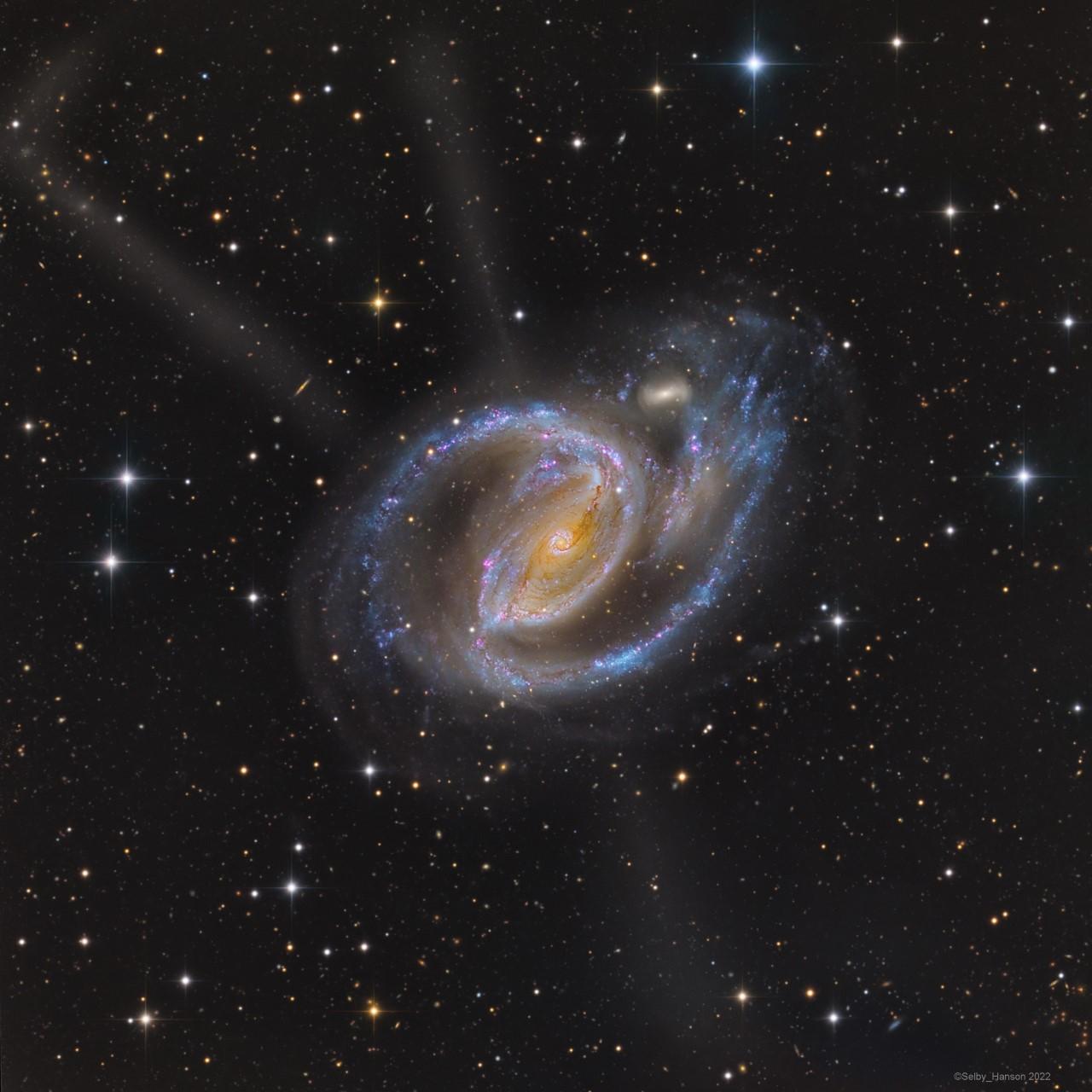 مجرّة حلزونيّة بنواتها المصفرّة  وذراعيها الحلزونيين الزرقاوين المرقّطين بالوردي. تظهر مجرّة صغيرة مصاحبة بجانبها وتدفّقات تبدو وكأنها تشع منها.