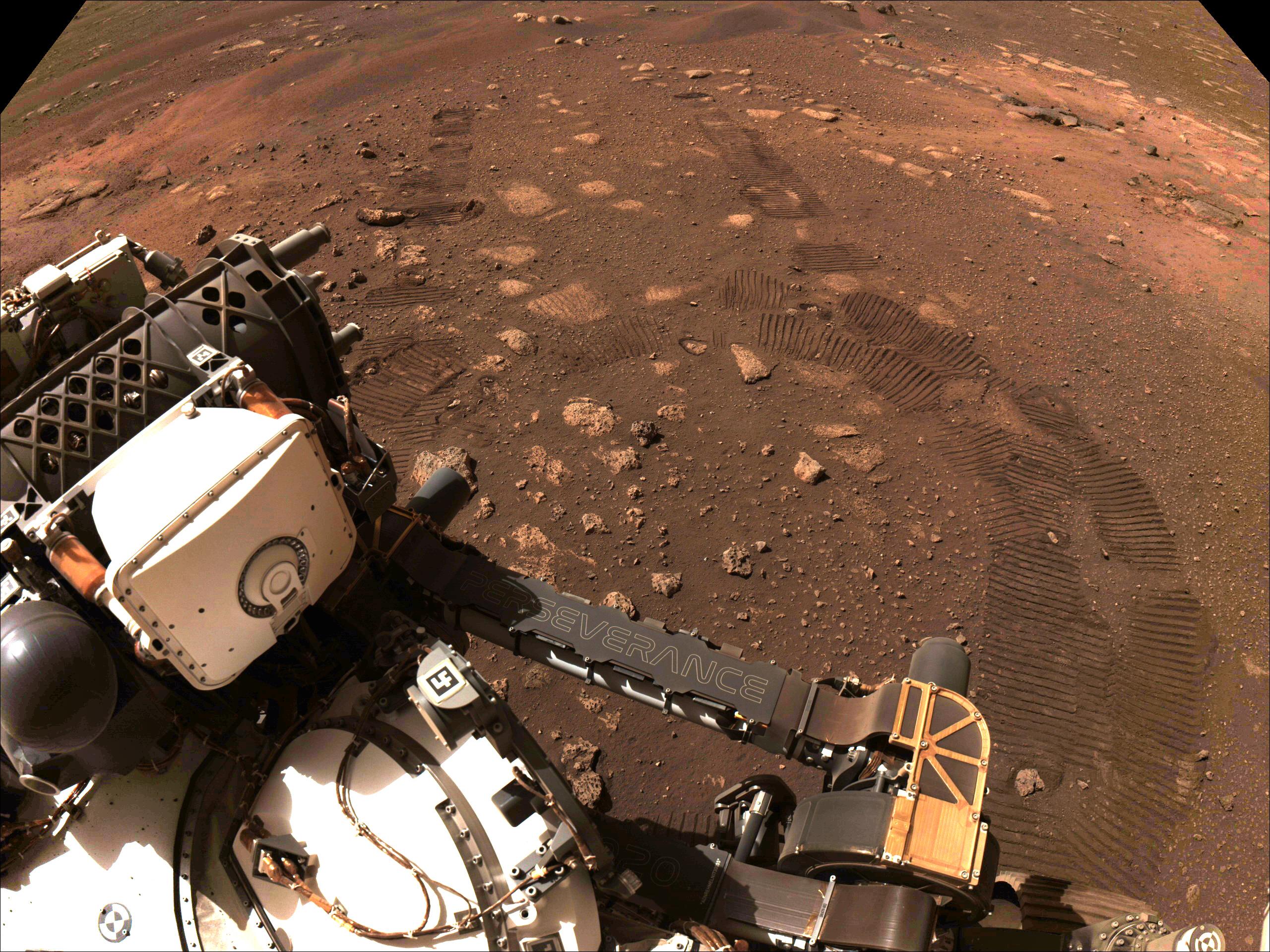 صورة يظهر في أسفلها جزء من عربة المُثابرة ومن خلفها سطح المريخ الذي تظهر على تربته آثار عجلاتها كخطين وقوس.