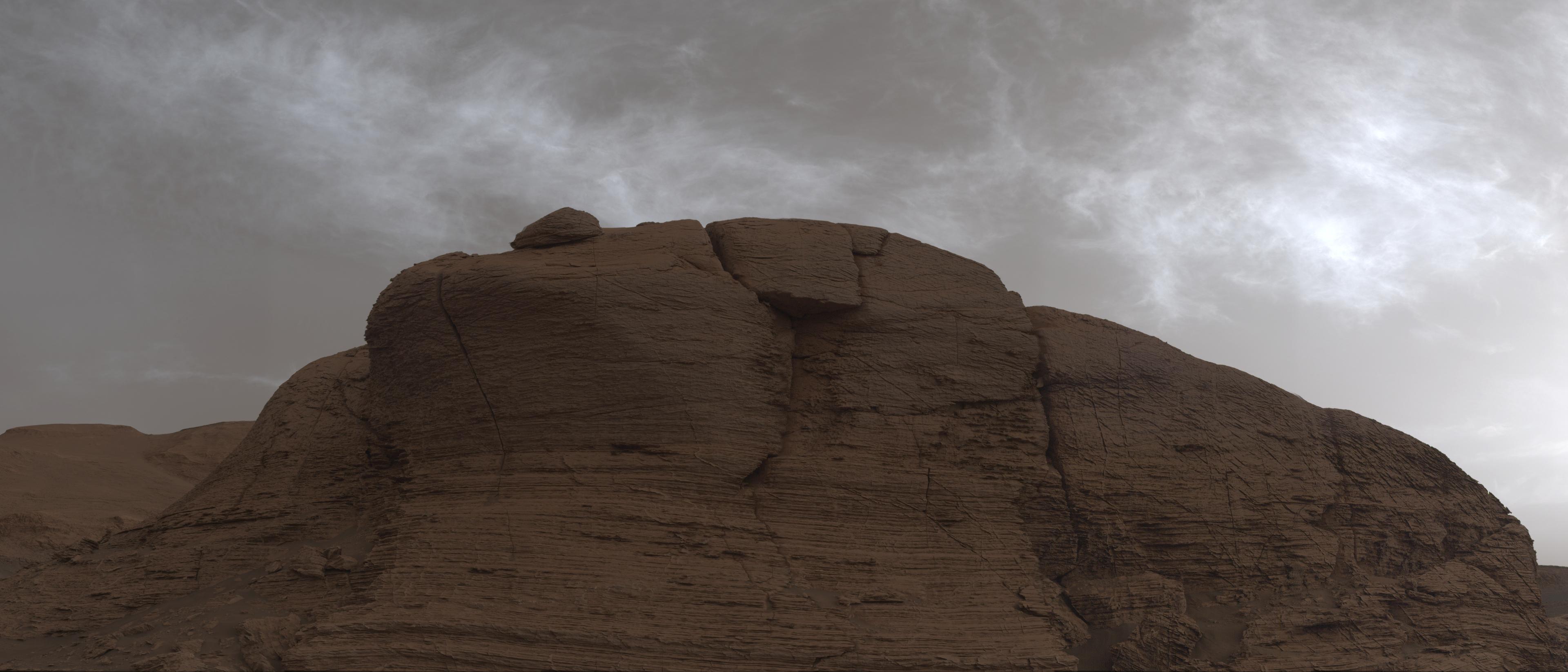 وجه جبل مرّيخي متعدّد الطبقات تظهر عليه آثار التجوية وتتألّق من فوقه السحب عاكسةً ضوء الشمس التي غابت للتو.