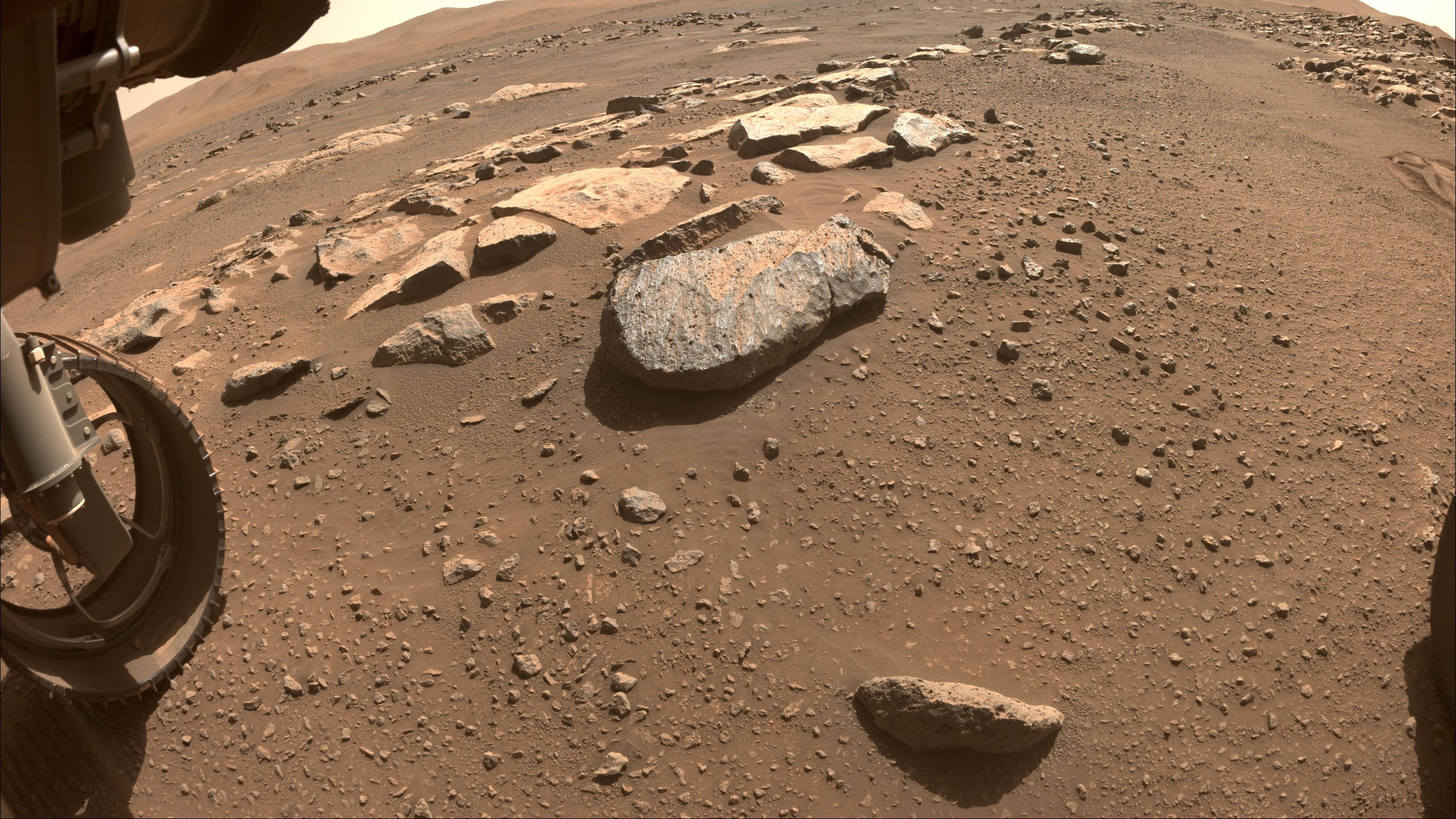 صورة لسطح المرّيخ المكتسي بالأتربة والأحجار وهناك صخرة كبيرة بارزة في وسط الصورة التي تظهر على يسارها عجلة مركبة المثابرة.