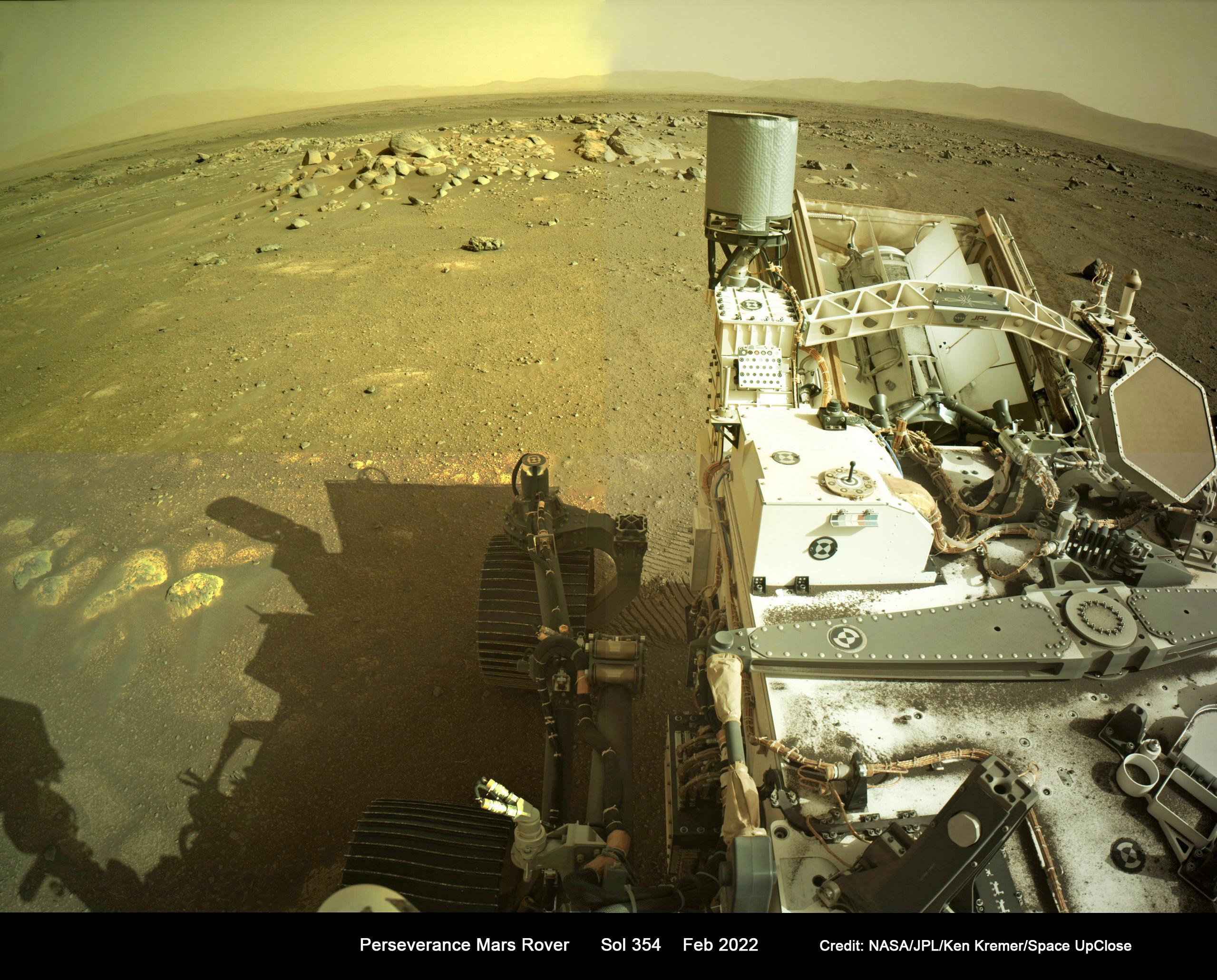 صورة بصبغة مصفرّة تظهر جزء من تجهيزات عربة المثابرة الجوّالة وبجانبها ظلها بينما تنبسط الأرض المريخية بصخورها وتلوح مرتفعات في البعيد
