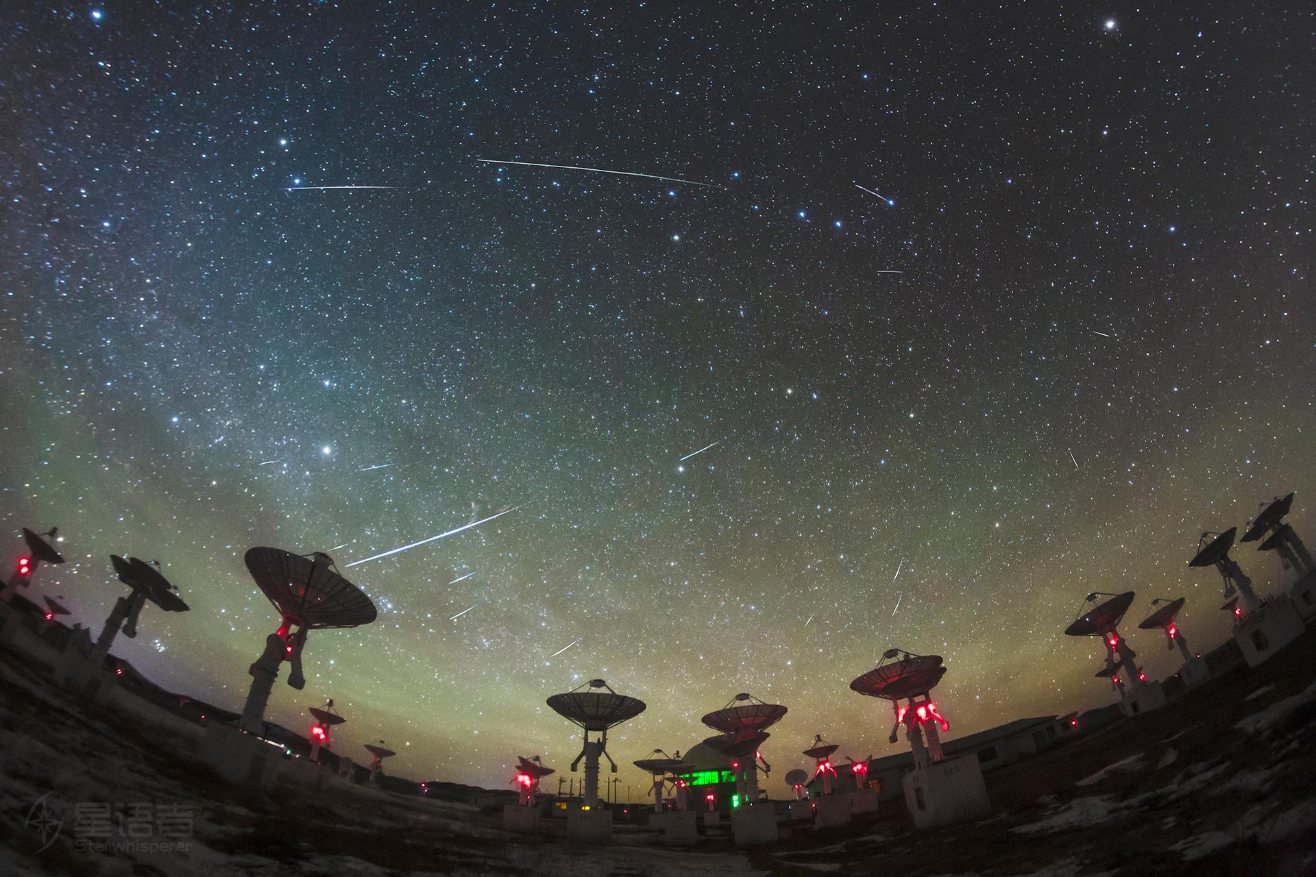 مصفوفة من صحون التلسكوبات الموجّهة نحو الأعلى وفوقها السماء المرصّعة بالنجوم يخطّ فيها عدد من الشهب