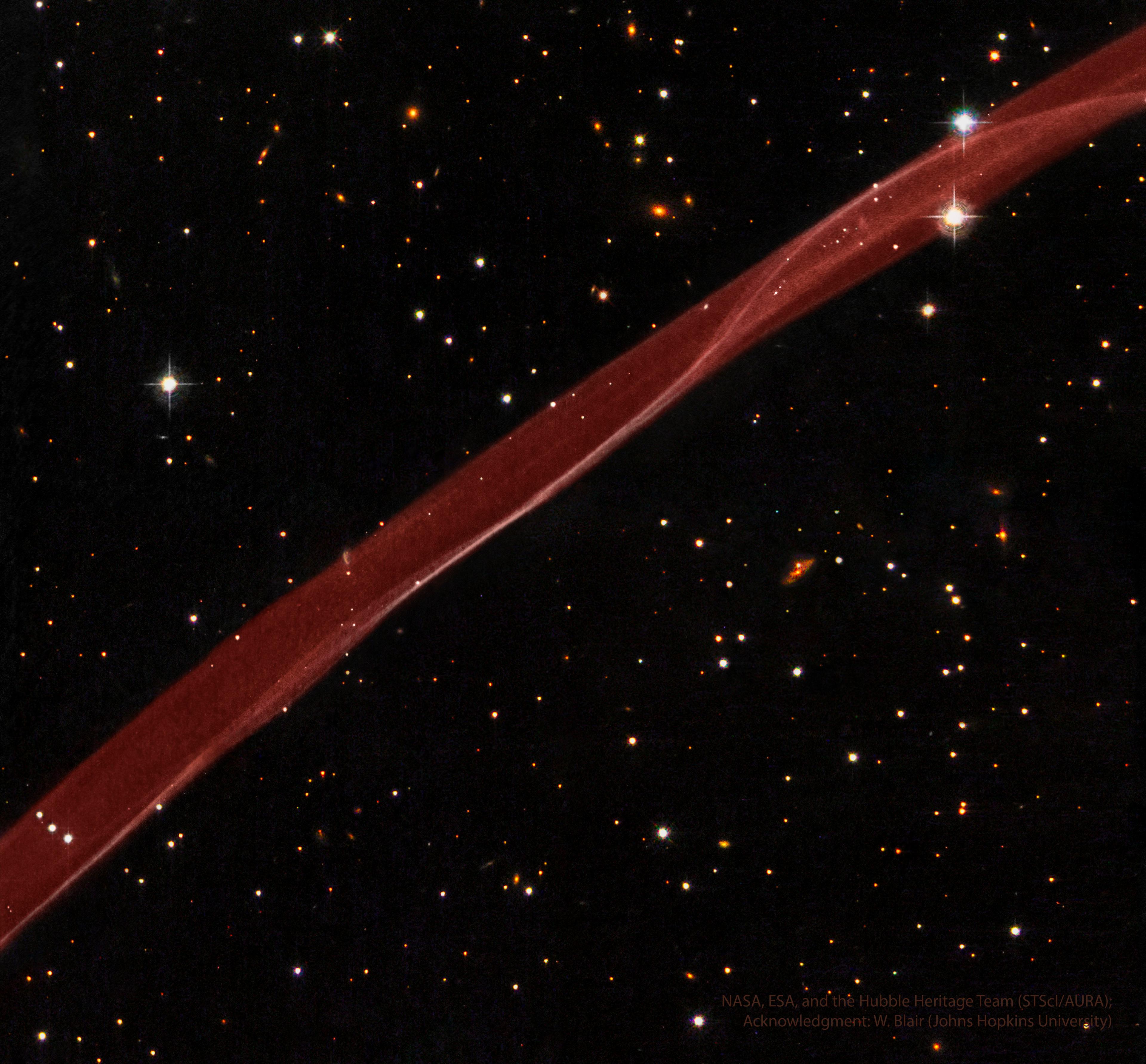 شريطةٌ سميكة شفّافة من الغاز الأحمر تجري من أسفل اليسار لأعلى اليمين. حقل نجومٍ مُظلم فيه نجومٌ ومجرّات يحيط بالشريطة الحمراء الساطعة.