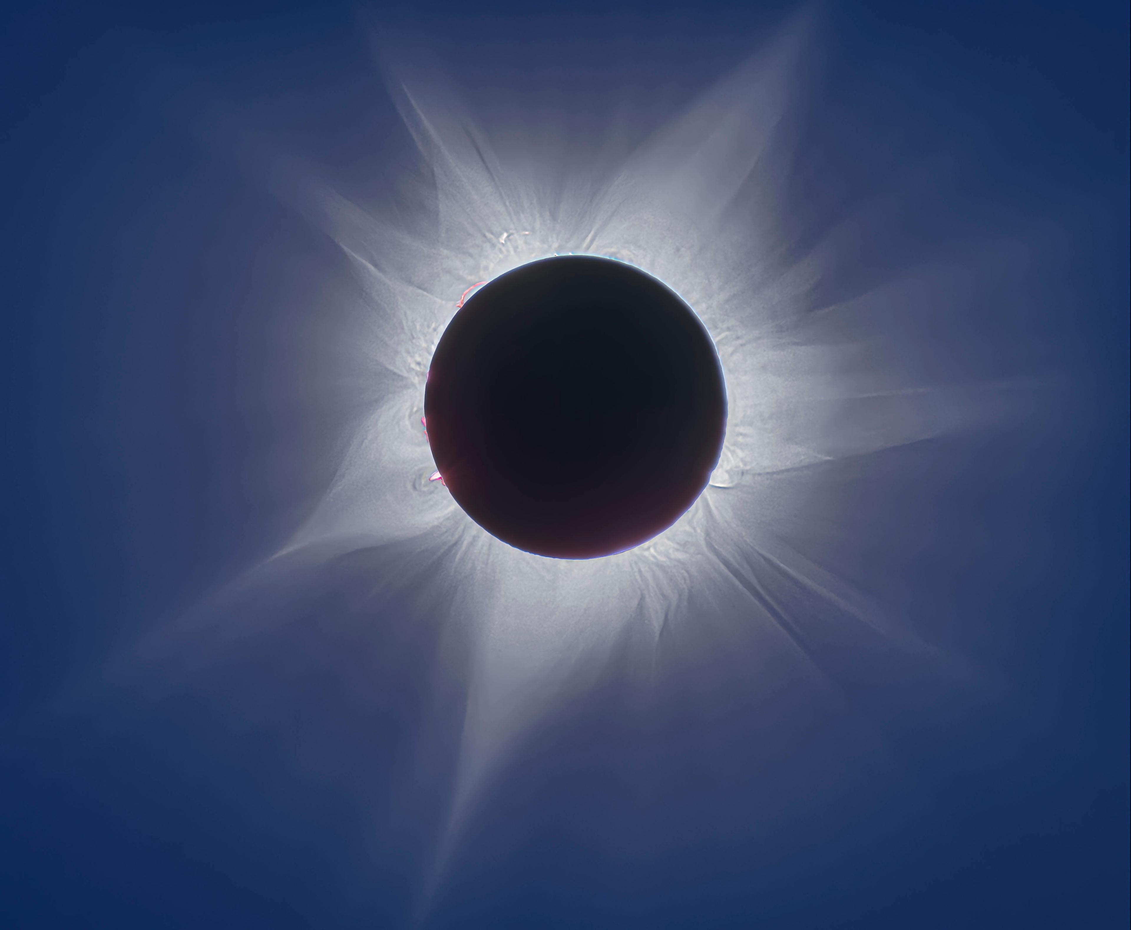 قرص أسود يمتدّ حوله الإكليل الشمسي كما تظهر بعض الشواظات الأكثف والأصغر، وحول ذلك كلّه السماء مزرقّة