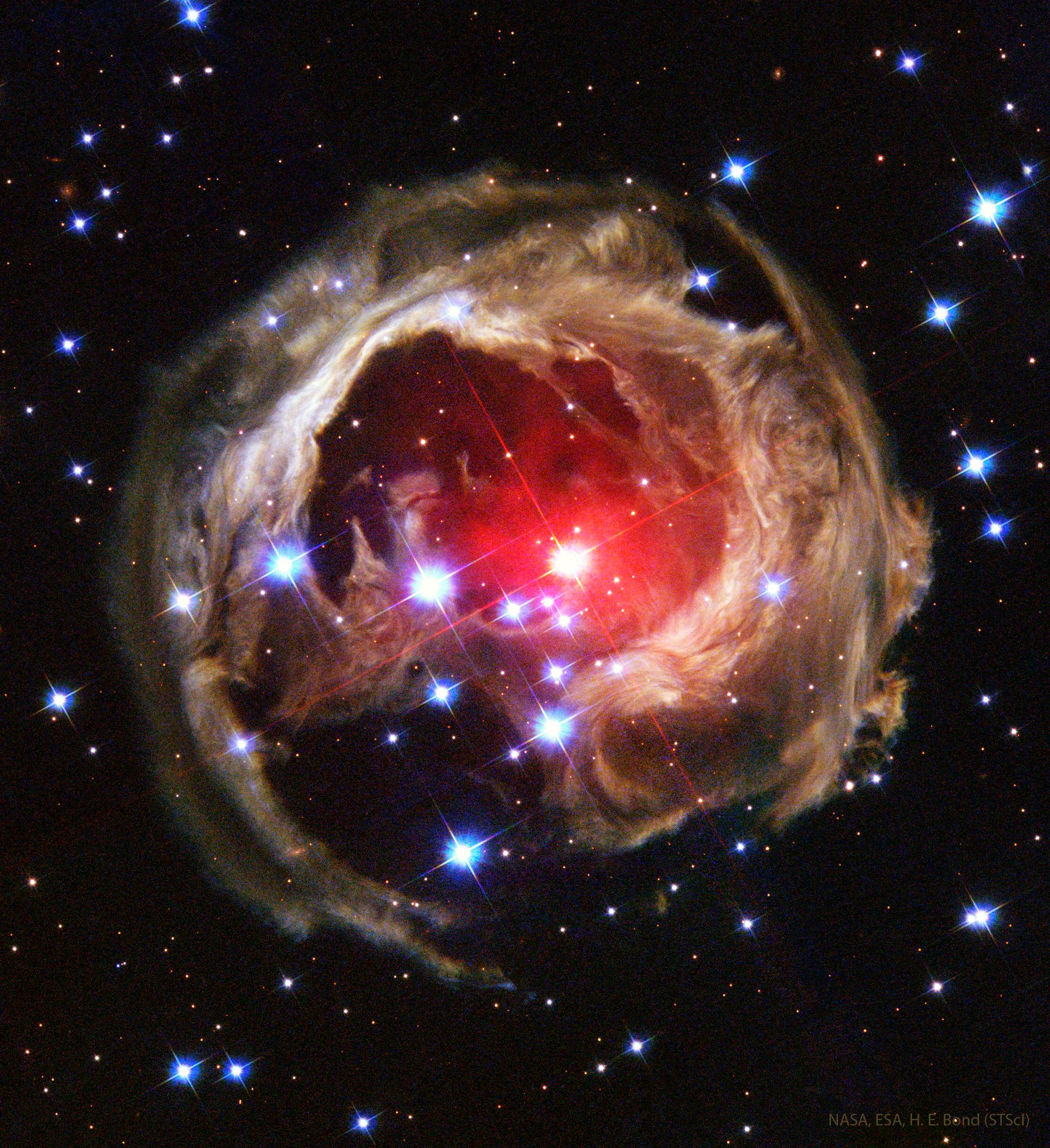 بُنية متوسّعة تظهر بلون محمر في الداخل وما يشبه طبقات من الألياف البنّية تحيط بها بينما تنتشر نجوم مدبّبة وأخرى صغيرة عبر الصورة