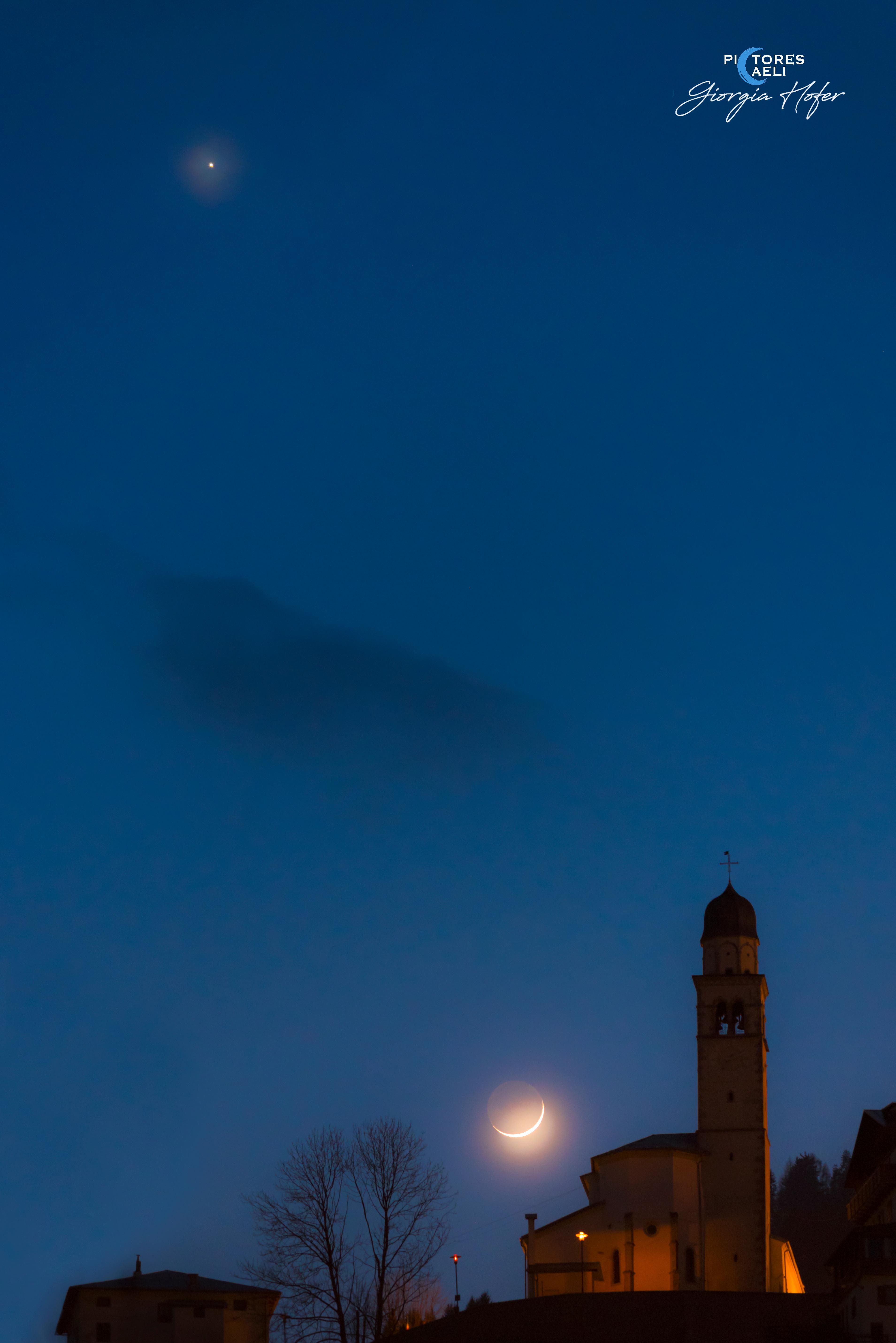 نجم الزُّهرة اللامع، والجانب الليليّ الخافت للقمر، والهلال الأهيَف المُنار بالشمس، متموضعين في سماء المساء إلى جانب برج كنيسة.