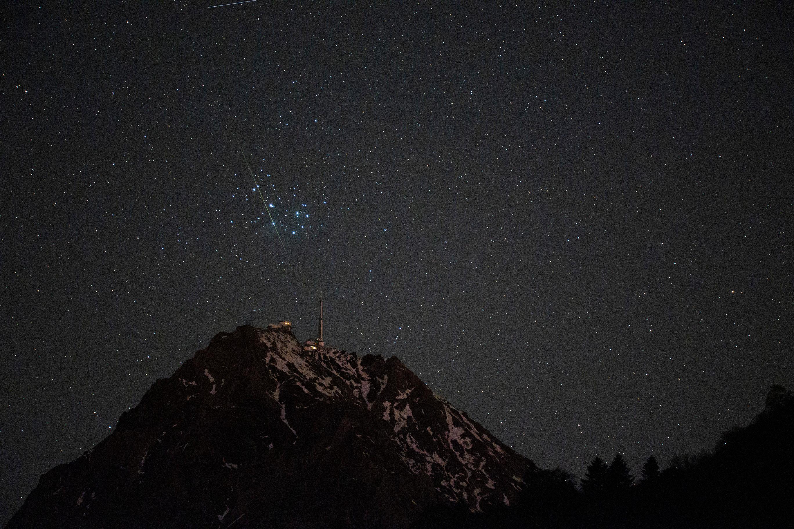 جبلٌ داكن مكسو بالثلوج وعلى قمّته بناءٌ يبدو كمنارة وفوقه شهابٌ يعبر أمام عنقود الثريّا على خلفيّة سماء ليل تتناثر فيها النجوم
