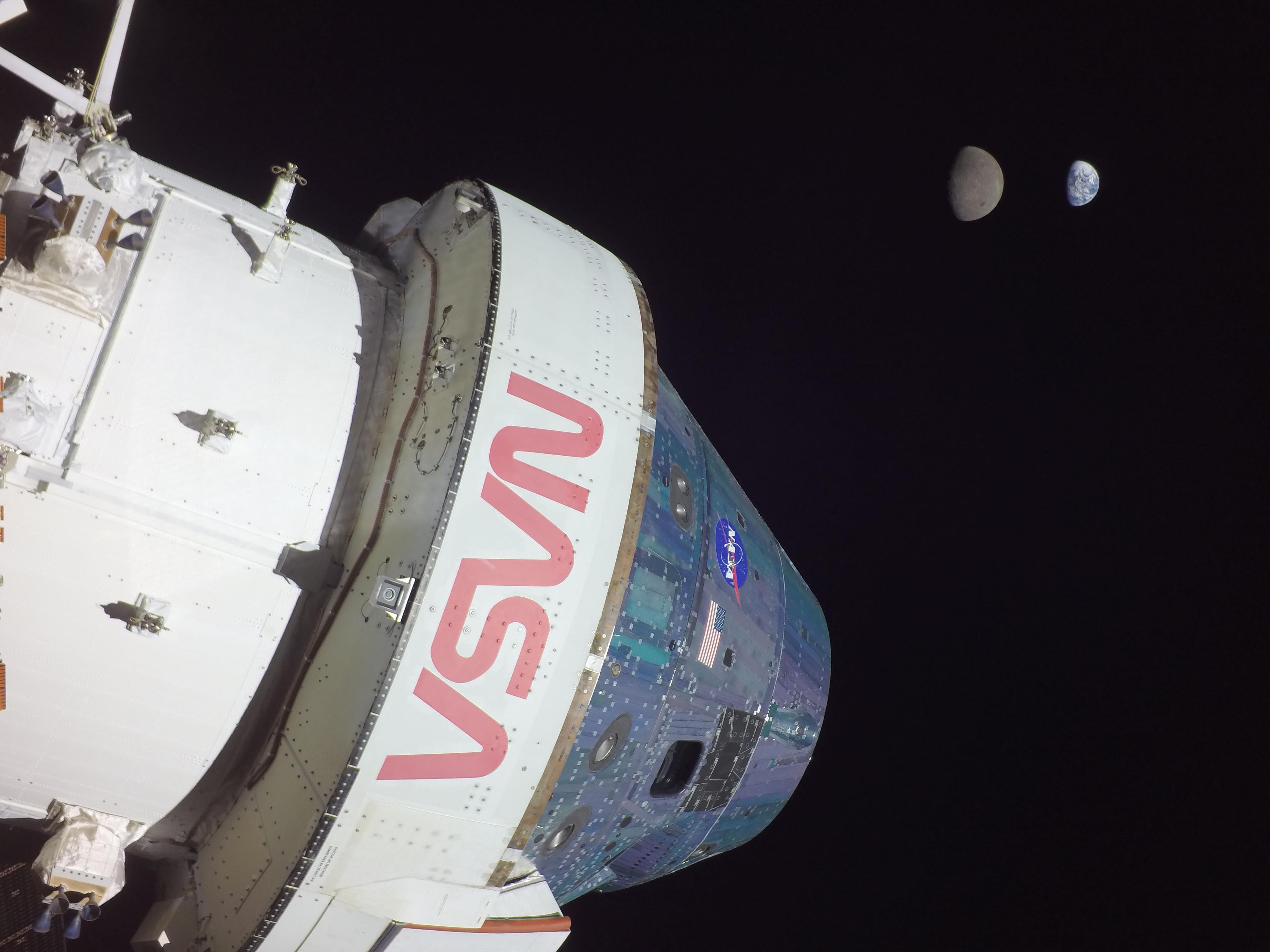 مركبة أورَيون تغطّي معظم الإطار بتفاصيل واضحة وعليها شعار ناسا، وفي البعيد يظهر القمر والأرض بطور الهلال الأحدب وبحجم ظاهري متقارب.