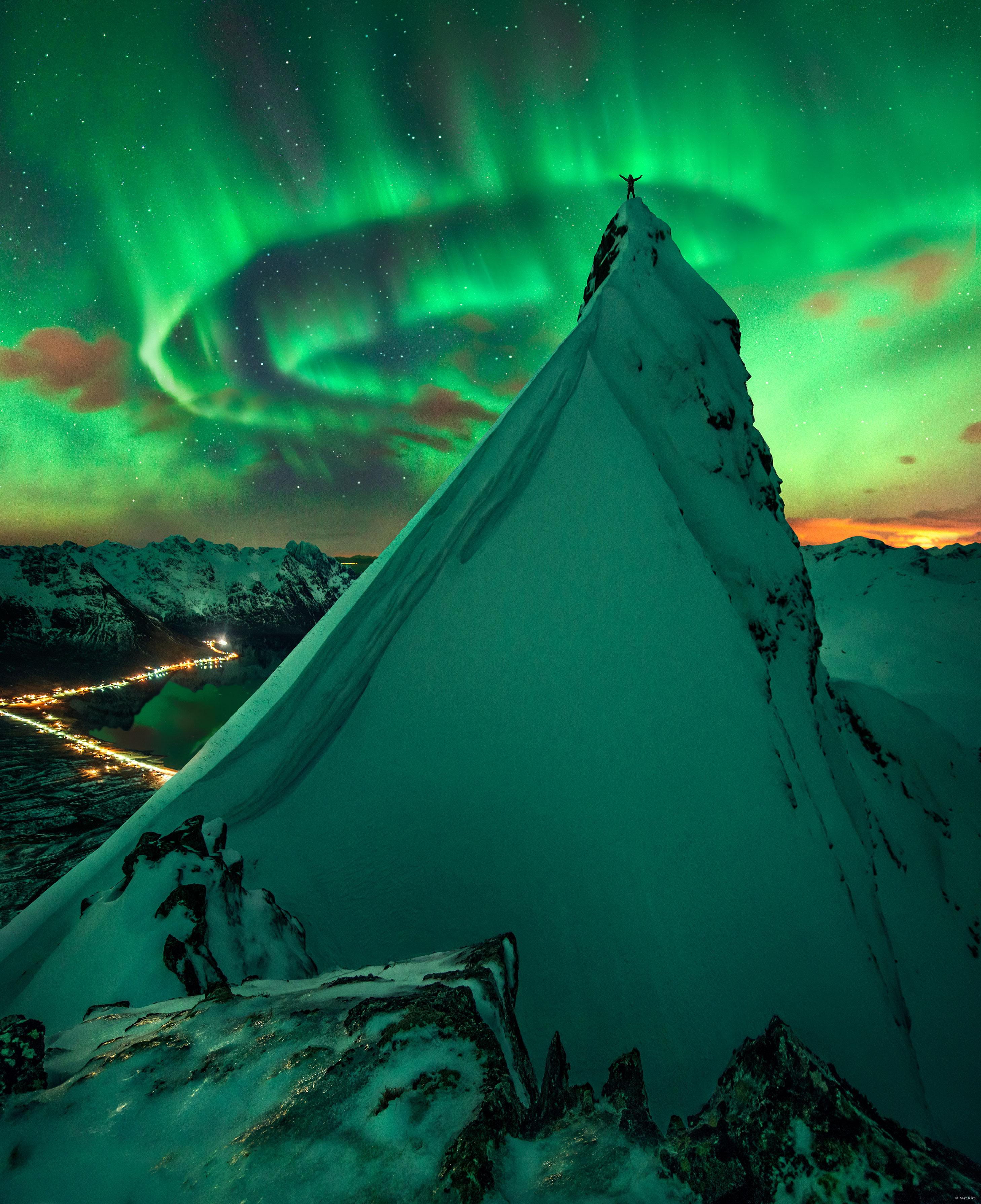 شخصٌ يقف فوق تلَّةٍ شديدة الانحدار مُغطّاة بالثلج رافعاً ذراعيه، بينما يُرى شفقٌ قطبيٌّ أخضر في المدى، وخلفه هناك نجومٌ مرئيّة.