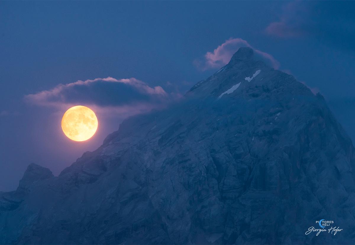 صورة لقمة جبل أنتيلاو والقمر شبه المكتمل يسطع قربه بينما يتشبّع الجبل والسماء بتدرجات اللون الأزرق بُعيد غروب الشمس.