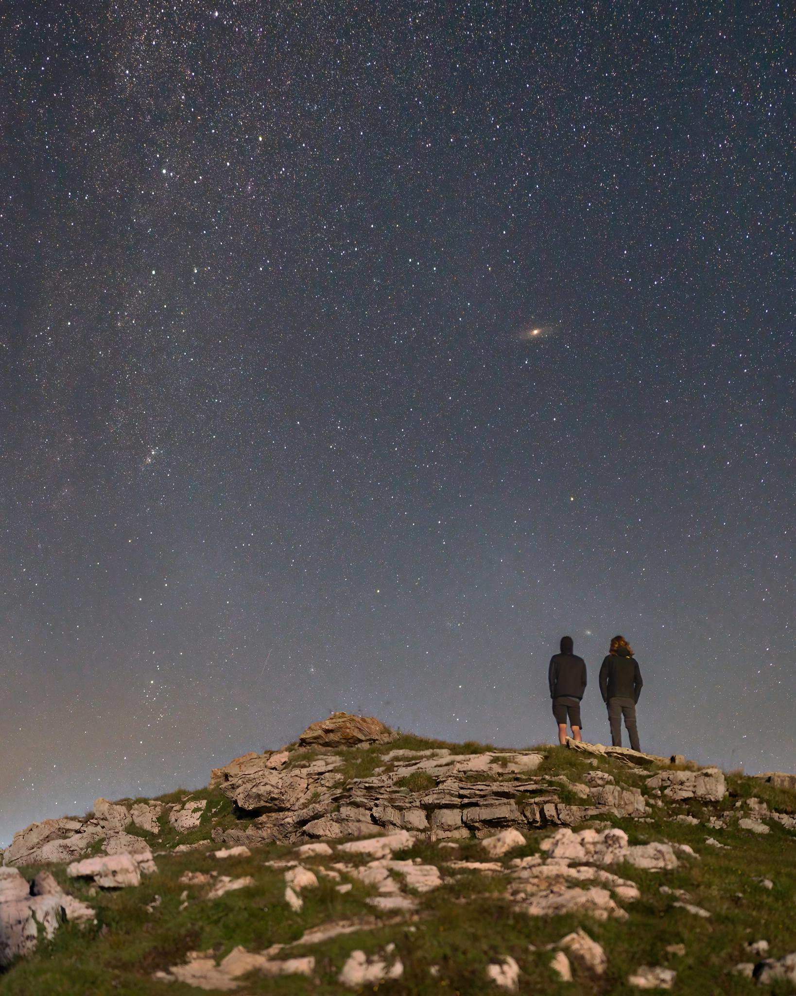 شخصان يقفان فوق تلّة بينما تسطع أمامهما نجوم الليل ويمتدّ مستوي درب التبّانة على طول يسار الصورة، وتظهر فوقهما مجرة أندروميدا بنواة ساطعة وبينهما مجرّة المثلّث.