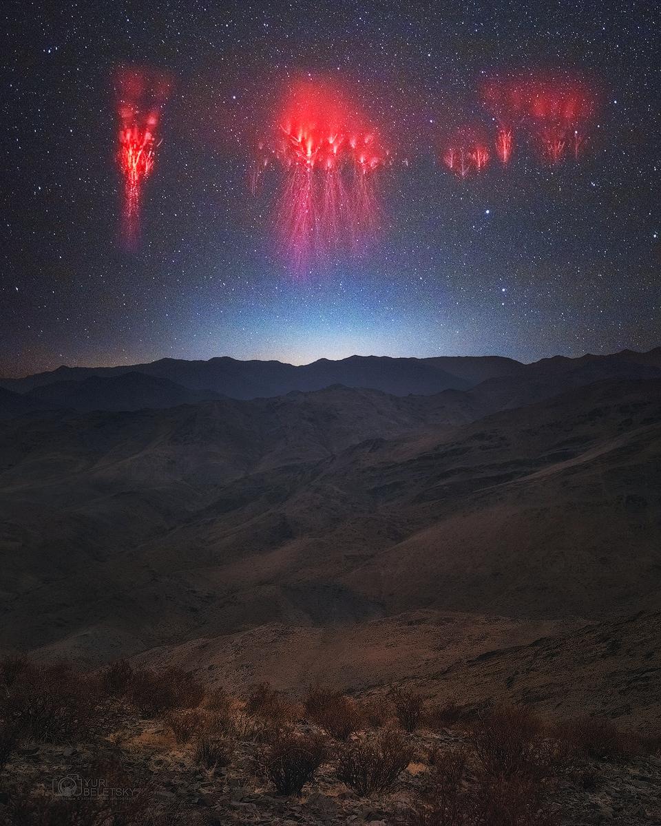 صورة تمتدّ فيها سلاسل الجبال باتجاه الأفق التي تظهر فوقه مجموعات من عفاريت البرق الحمراء على امتداد عرض الصورة تقريباً ومن خلفها سماء الليل المرصّعة بالنجوم. يظهر وهجٌ مزرقٌّ ساطع في منتصف الأفق.