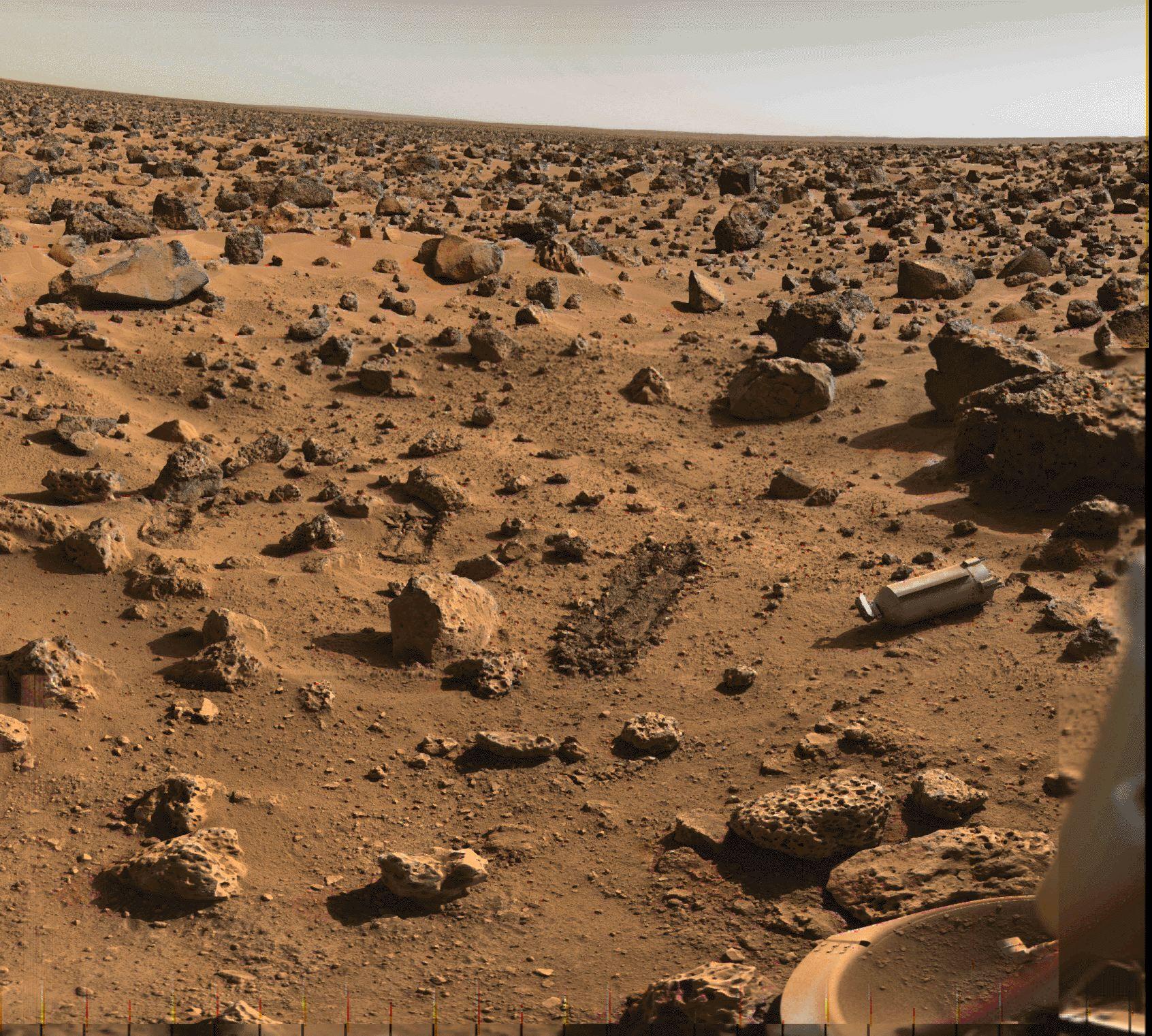 سطح المريخ في منطقة مليئة بالصخور والجلاميد التي تمتدّ للأفق. يظهر في وسط الصورة خندقان صغيران، وعلى اليمين الغطاء الواقي المقذوف من مركبة ڤايكينگ، إضافة إلى أحد مساند أقدام الهابط المغطىّ بالغبار في أسفل يمين الصورة.